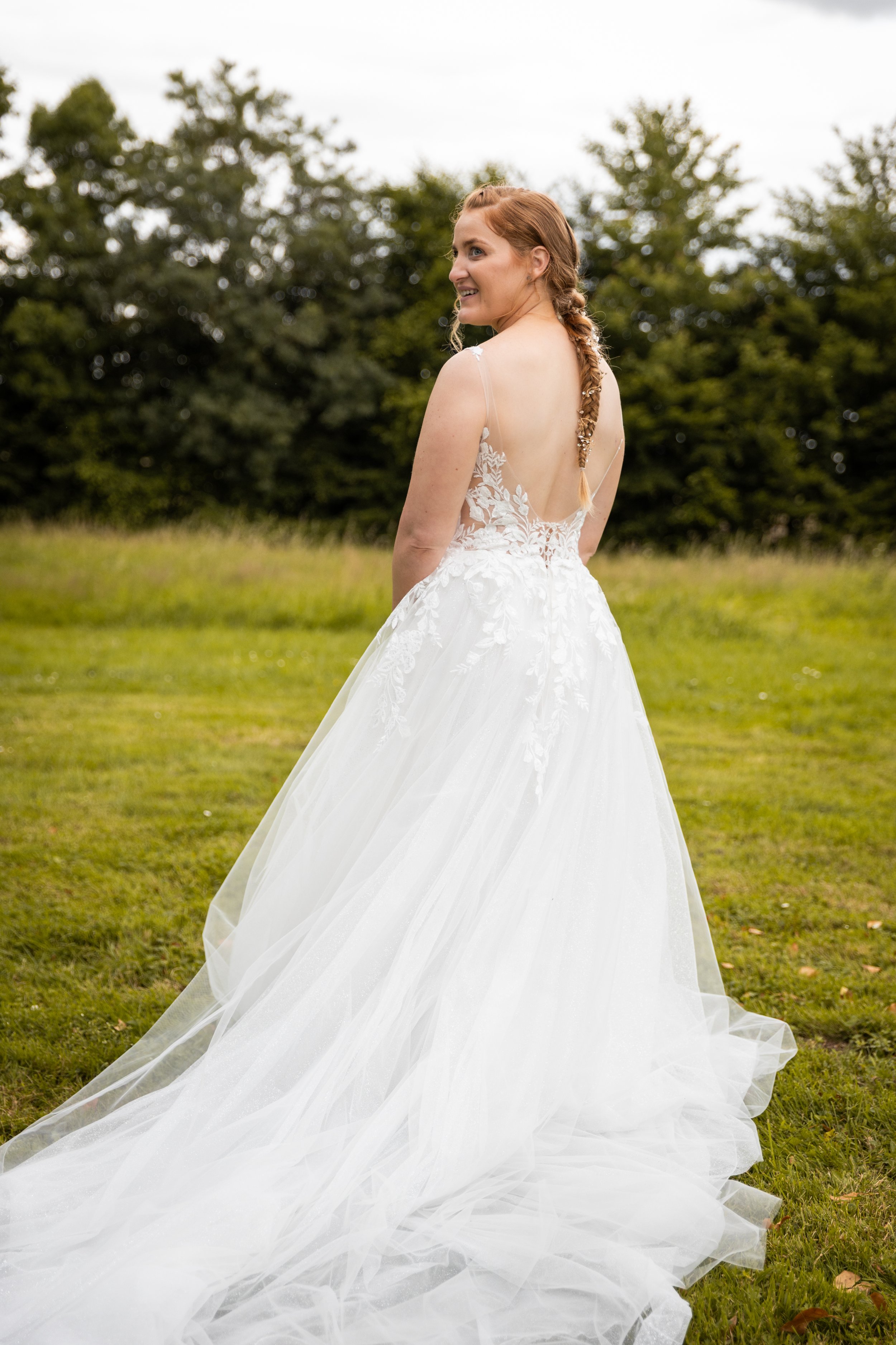 Une femme en robe de mariée blanche dans un champ vert, regardant par-dessus son épaule avec un sourire, ses cheveux roux attachés en tresse.