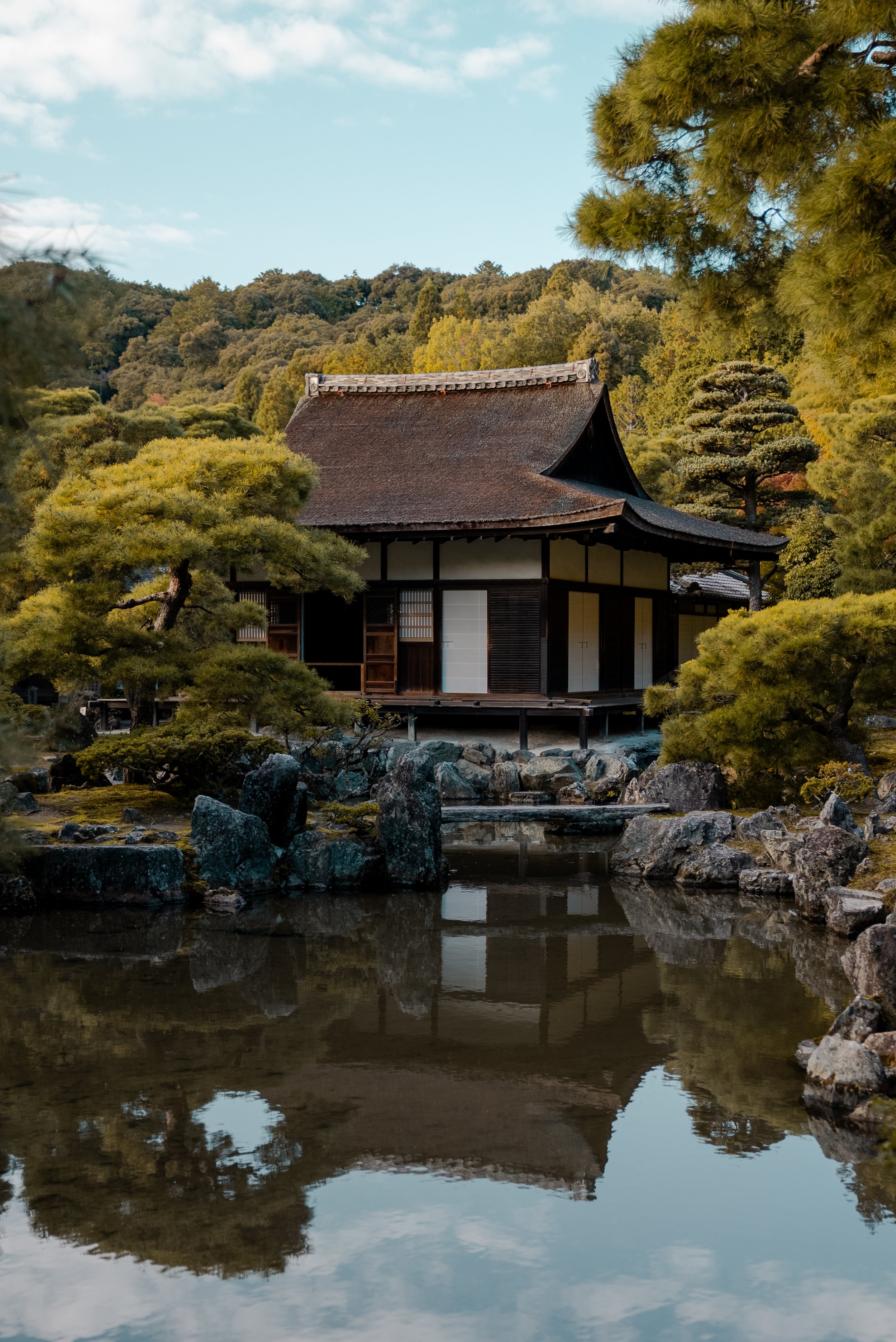 Une maison traditionnelle japonaise en bois avec un toit en tuiles, entourée d'arbres dans un jardin japonais avec un étang en reflection.