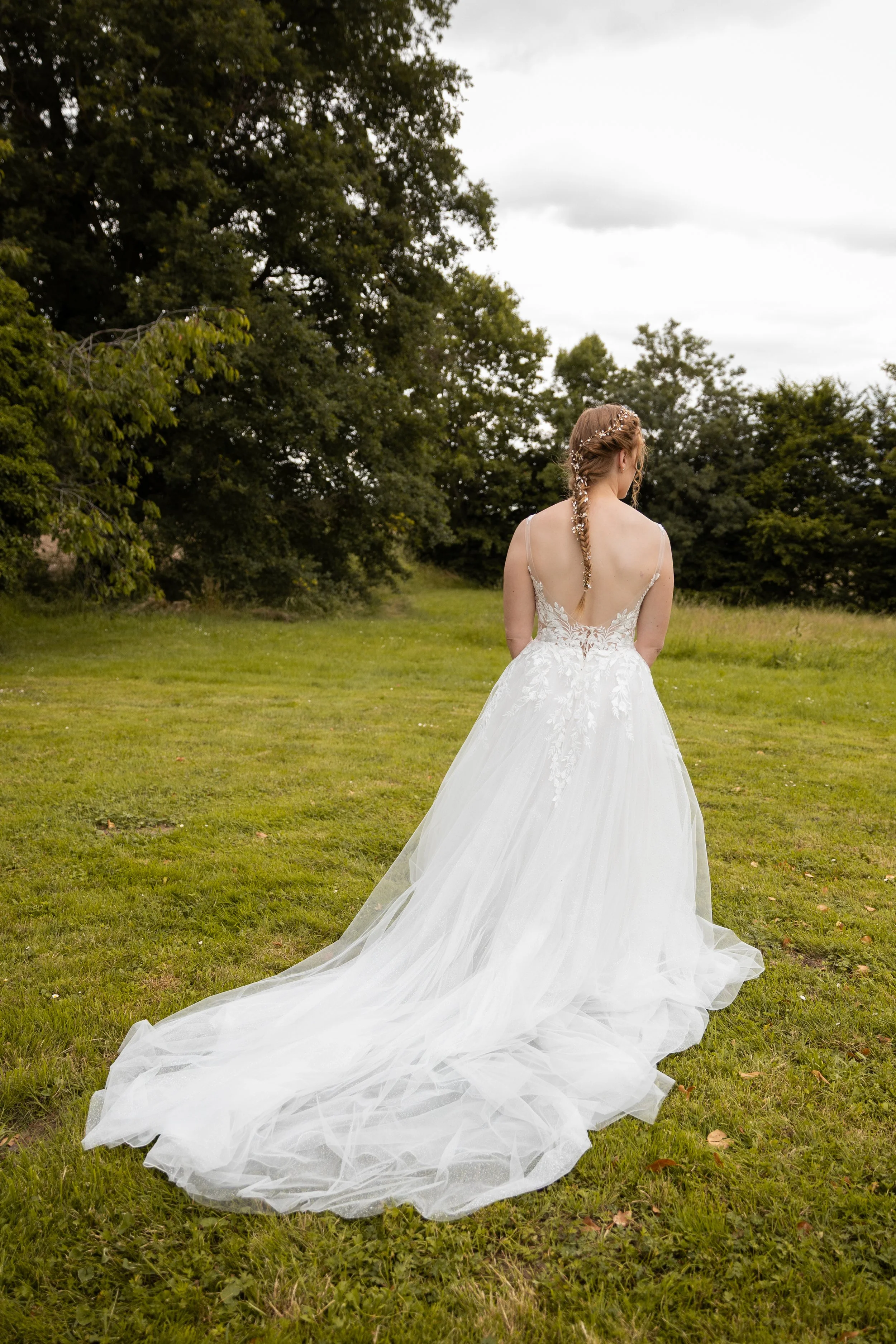 Une femme en robe de mariée blanche, vue de dos, dans un champ vert avec des arbres en arrière-plan, un ciel nuageux.