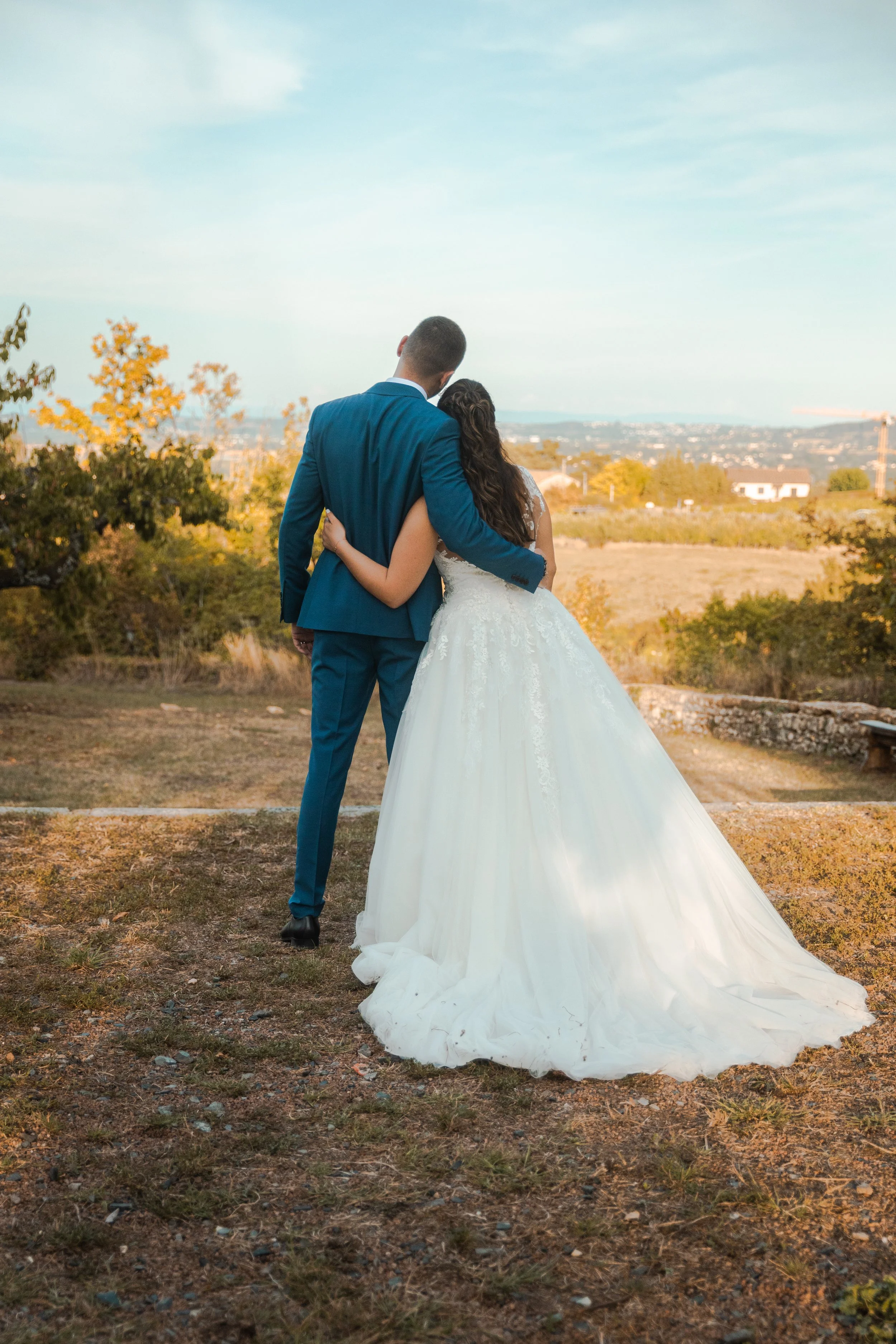 Un couple de mariés en tenue de mariage se tient côte à côte dans un paysage en plein air lors du coucher de soleil.