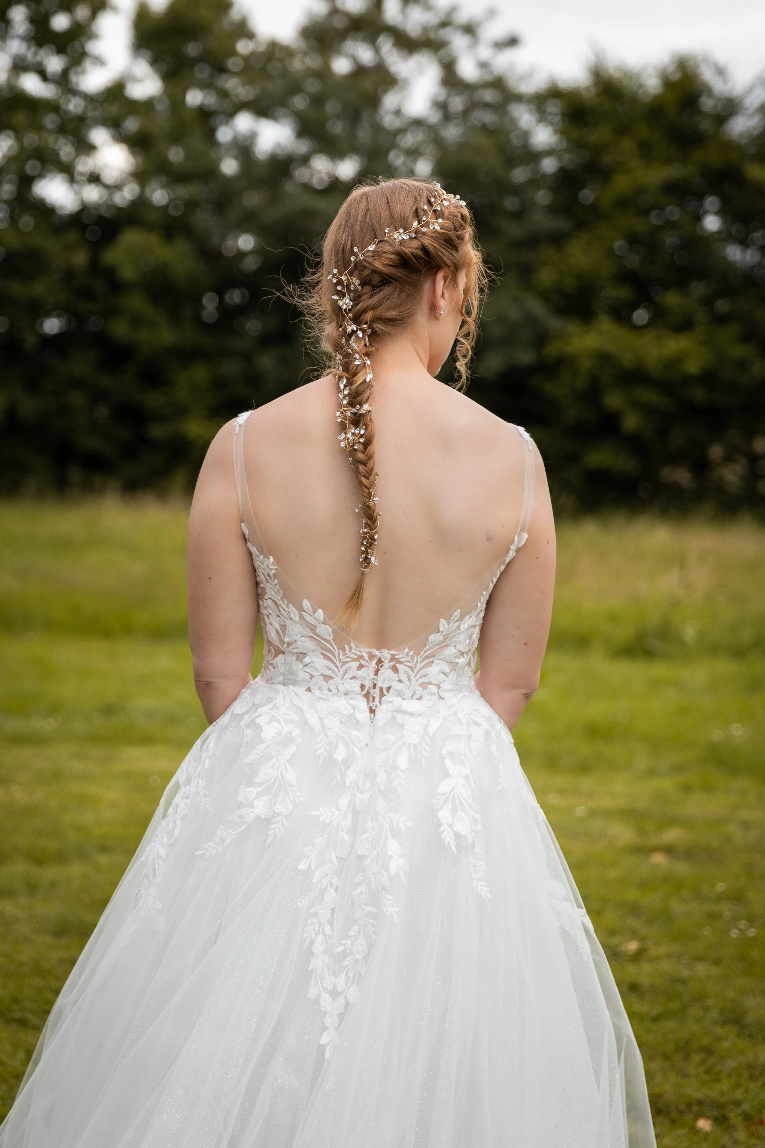 Une femme en robe de mariage blanche avec des détails de dentelle travaillée sur le dos, vue de dos, avec une coiffure tressée ornée de bijoux, dans un espace extérieur avec de l'herbe et des arbres.