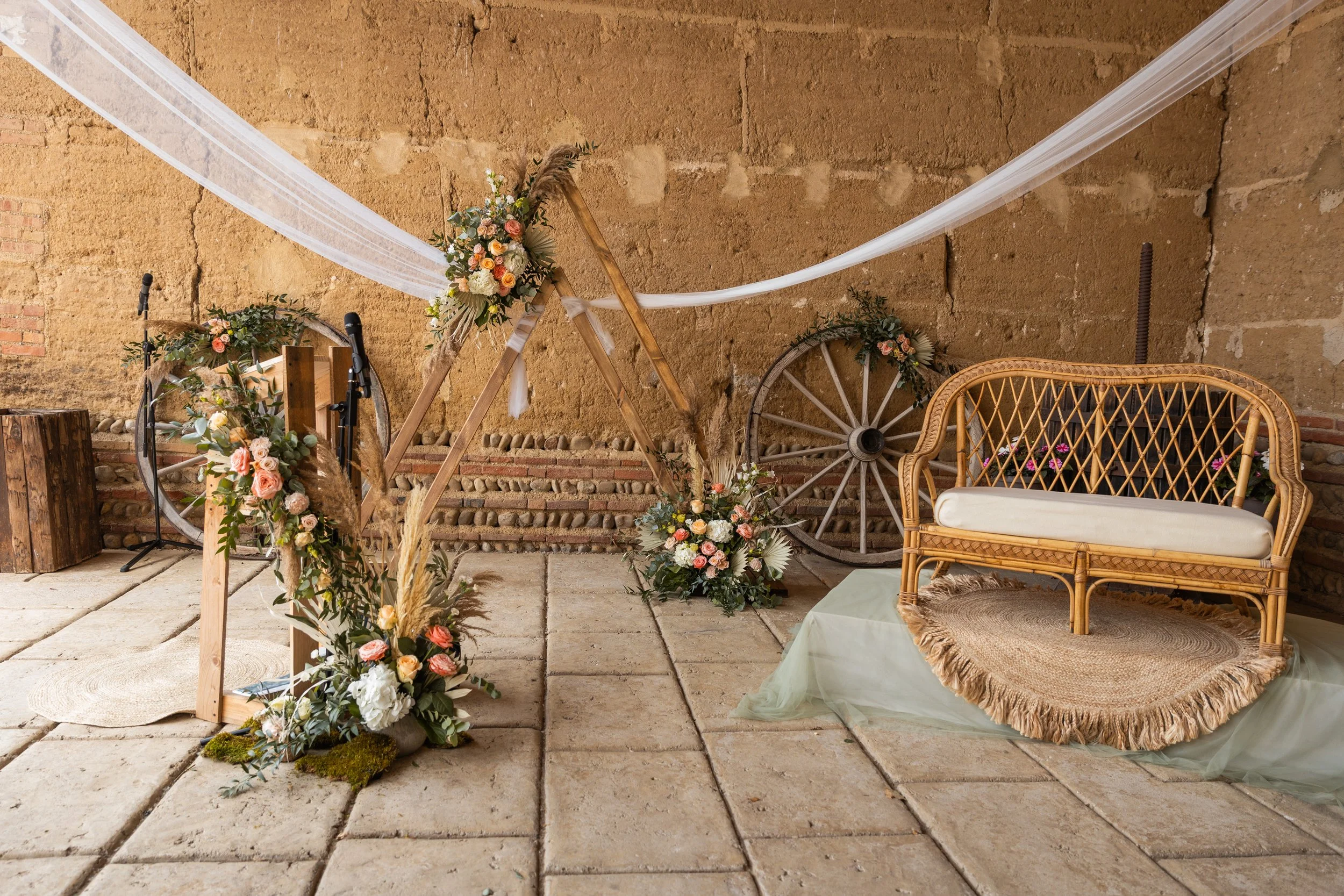 Décoration de mariage avec un cercle en bois orné de fleurs, deux roues en bois appuyées contre un mur en pierre, un banc en rotin avec coussin blanc et un tapis rond en jute.