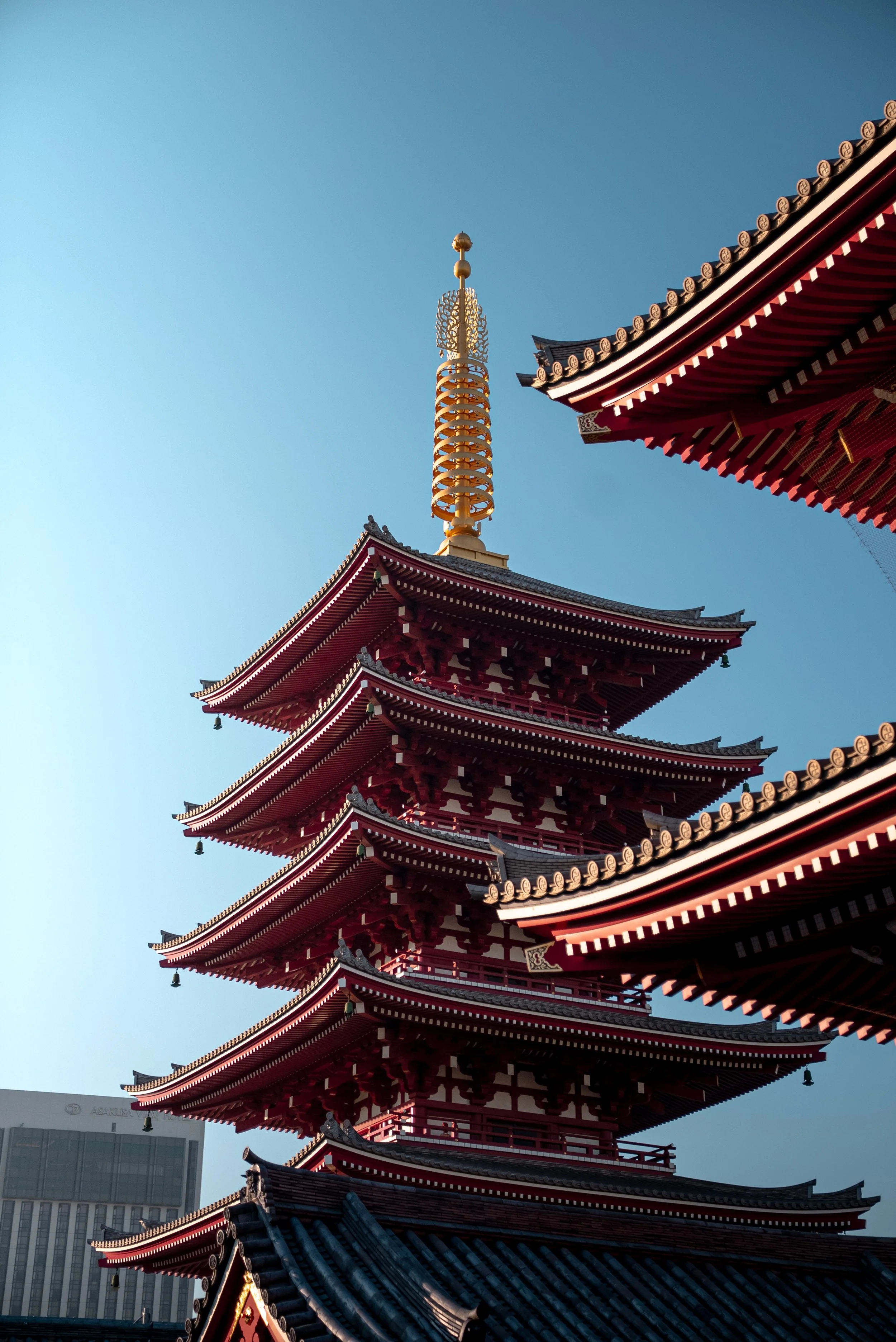 Une pagode japonaise à plusieurs étages avec un toit en tuiles noires et une tour dorée au sommet contre un ciel bleu.