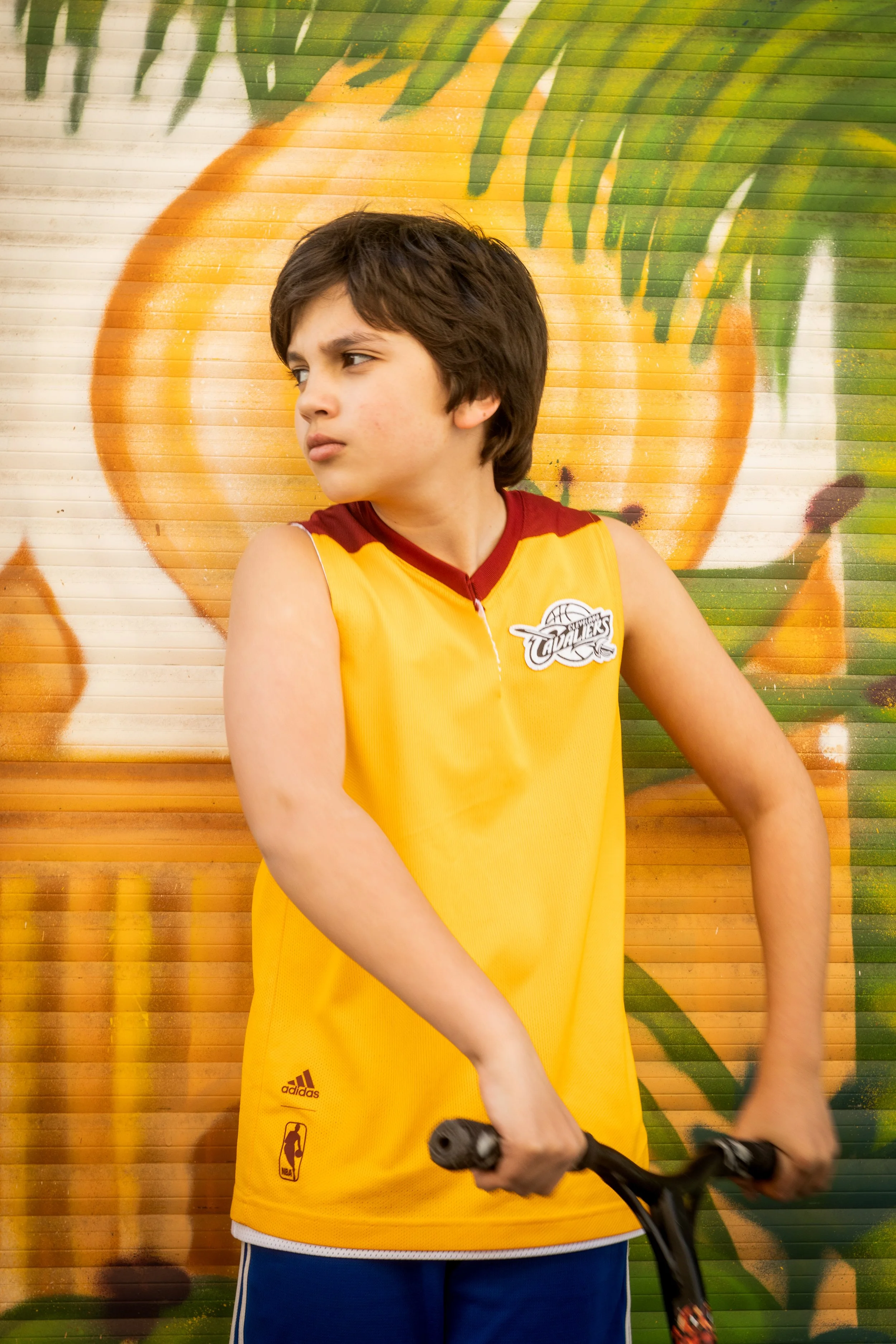 Jeune garçon portant un maillot de basketball jaune avec des détails rouges, tenant un hoverboard, se tient devant un mur peint avec une grande image d'une orange.