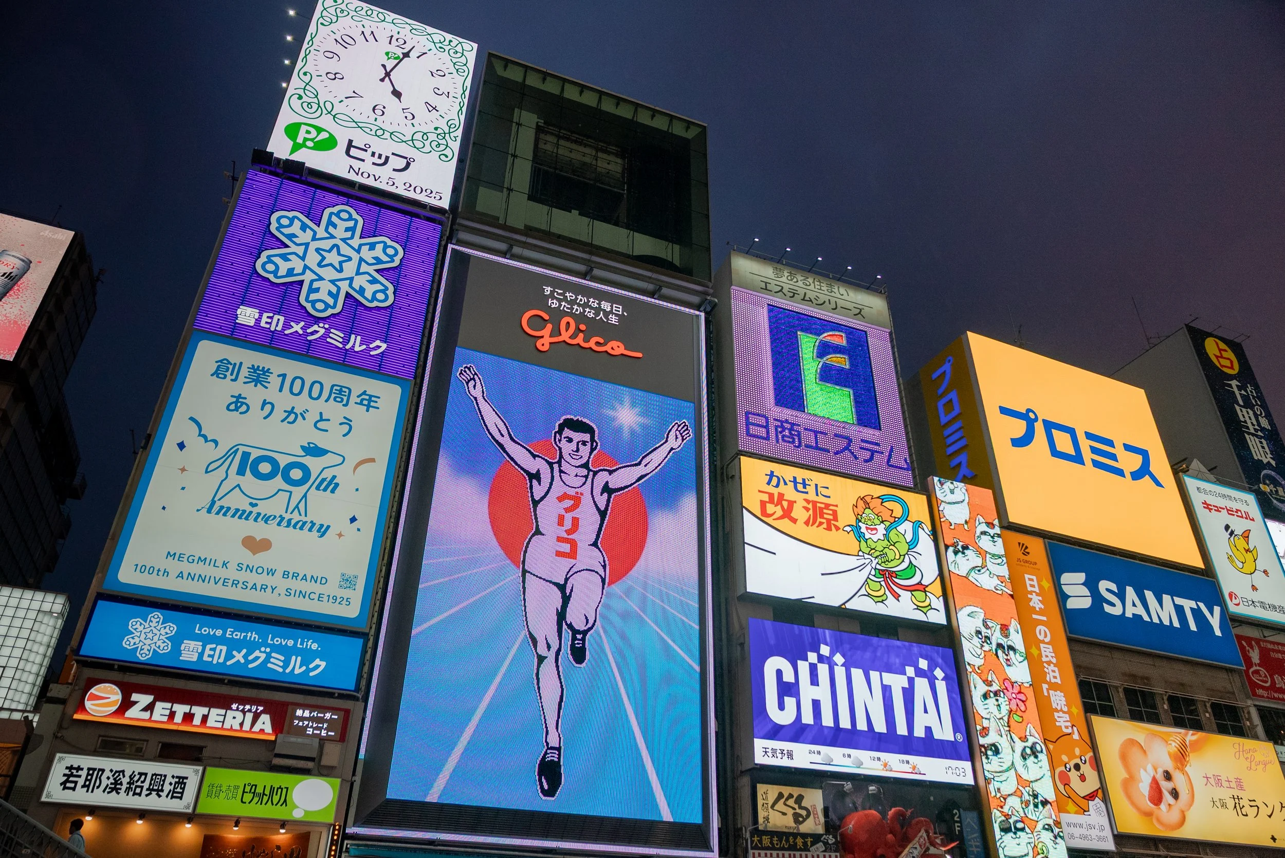 Écrans publicitaires illuminés dans un quartier de ville la nuit, avec un compte à rebours, un dessin de marathonien et diverses publicités en japonais.