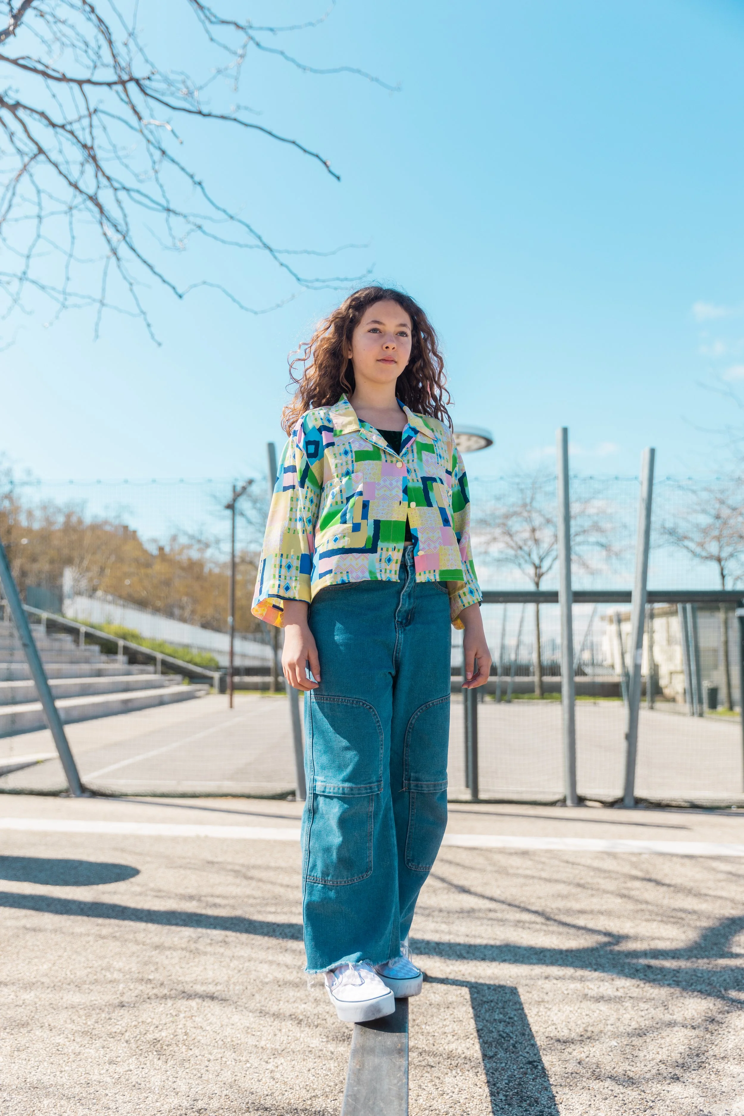 Jeune femme debout sur un équilibreur en extérieur, portant une chemise colorée et un jeans, avec un terrain de basketball en arrière-plan et un ciel bleu.