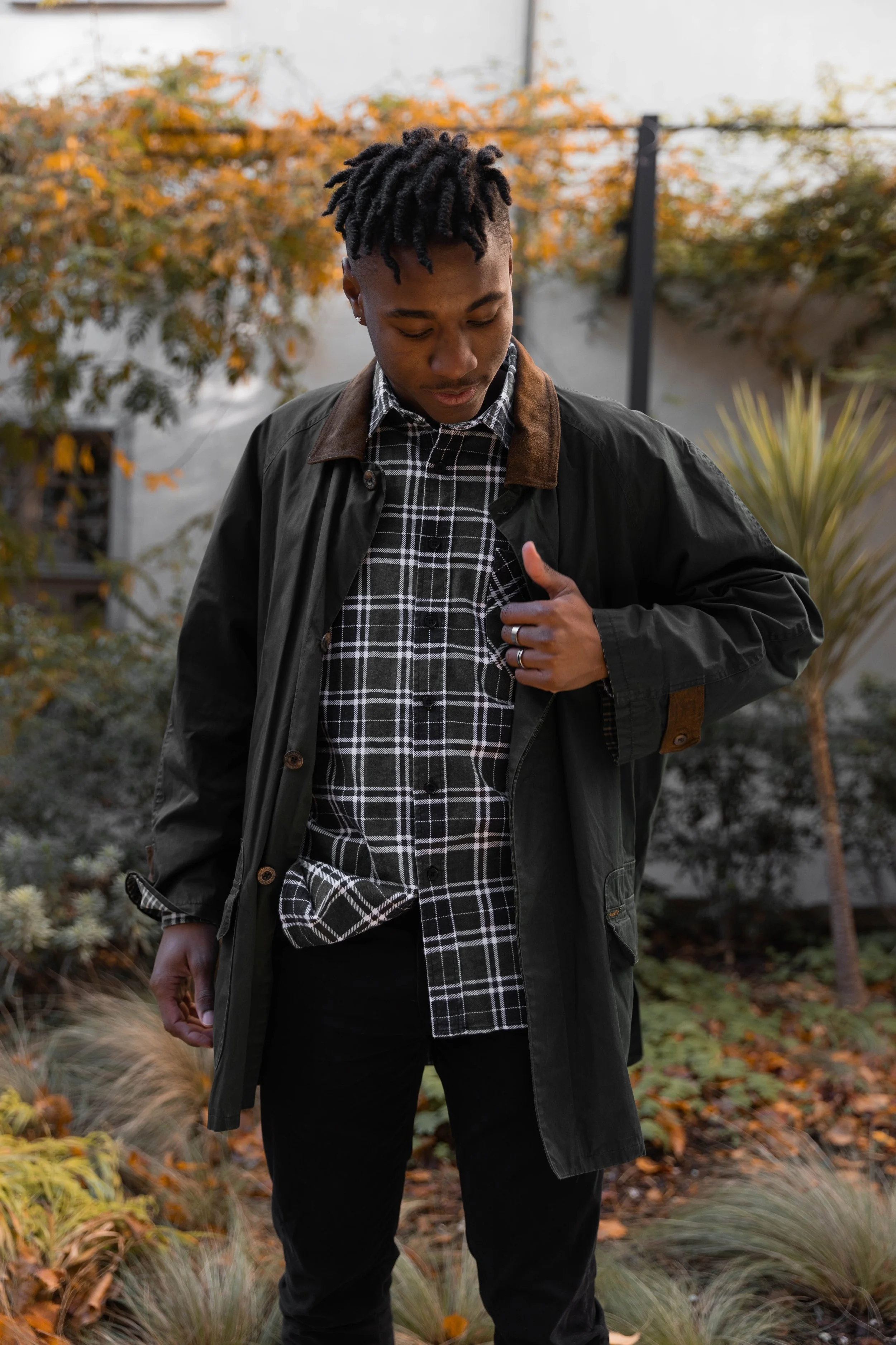 Un jeune homme regarde vers le bas, portant une veste noire et un chemisier à carreaux noirs et blancs, dans un jardin aux feuillages d'automne.