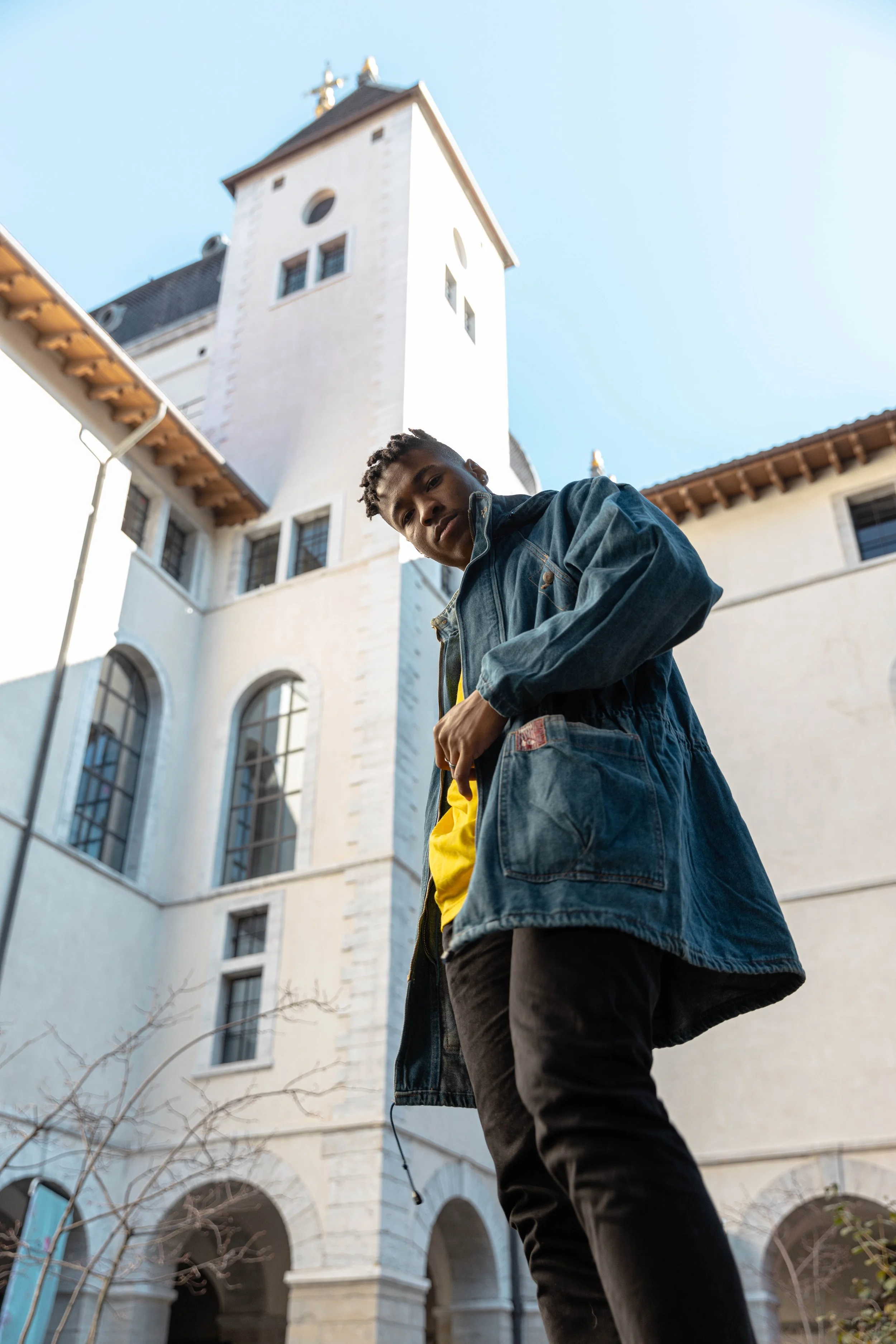 Jeune homme debout devant une vieille bâtisse en pierre avec un clocher à l'arrière-plan, portant une veste en denim jaune et sombre, regardant vers le bas.