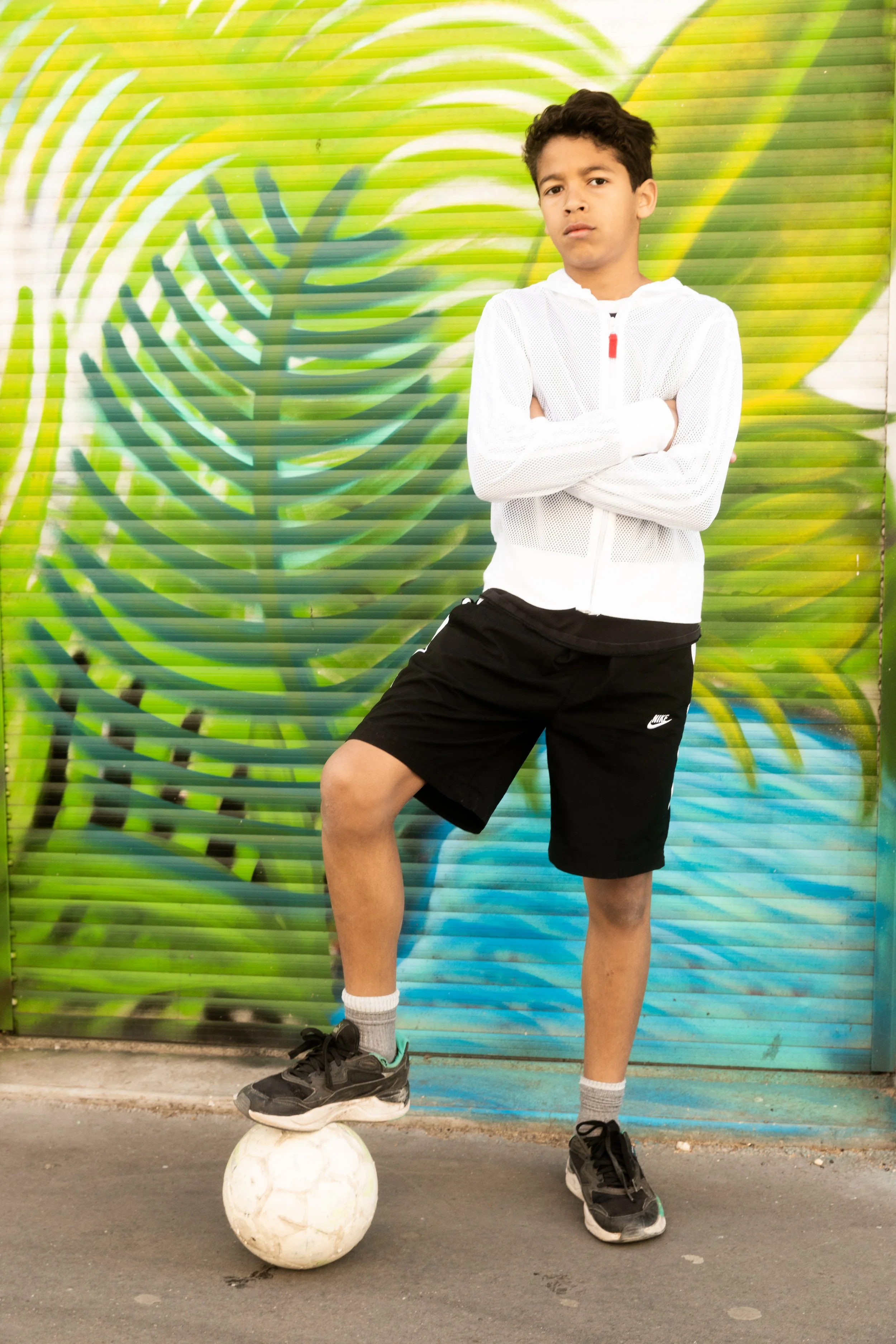 Un garçon debout avec bras croisés, portant un sweat blanc, un short noir Nike, et des baskets noires, avec un soccer sous le pied, en face d'un mur peint avec un motif de feuilles vertes.