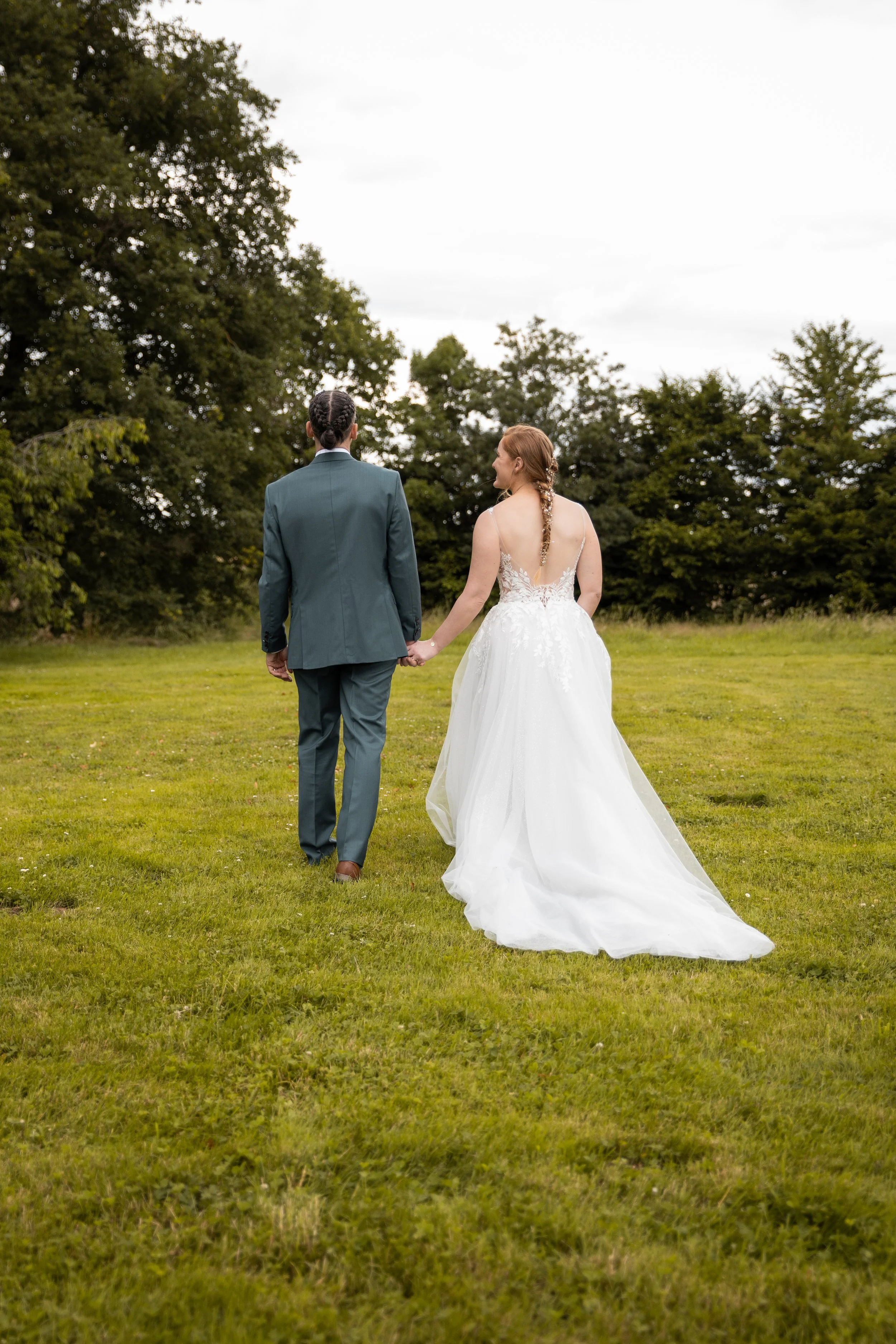 Jeune couple de mariés marchant main dans la main dans un champ, vu de dos, avec des arbres en arrière-plan, le jour.