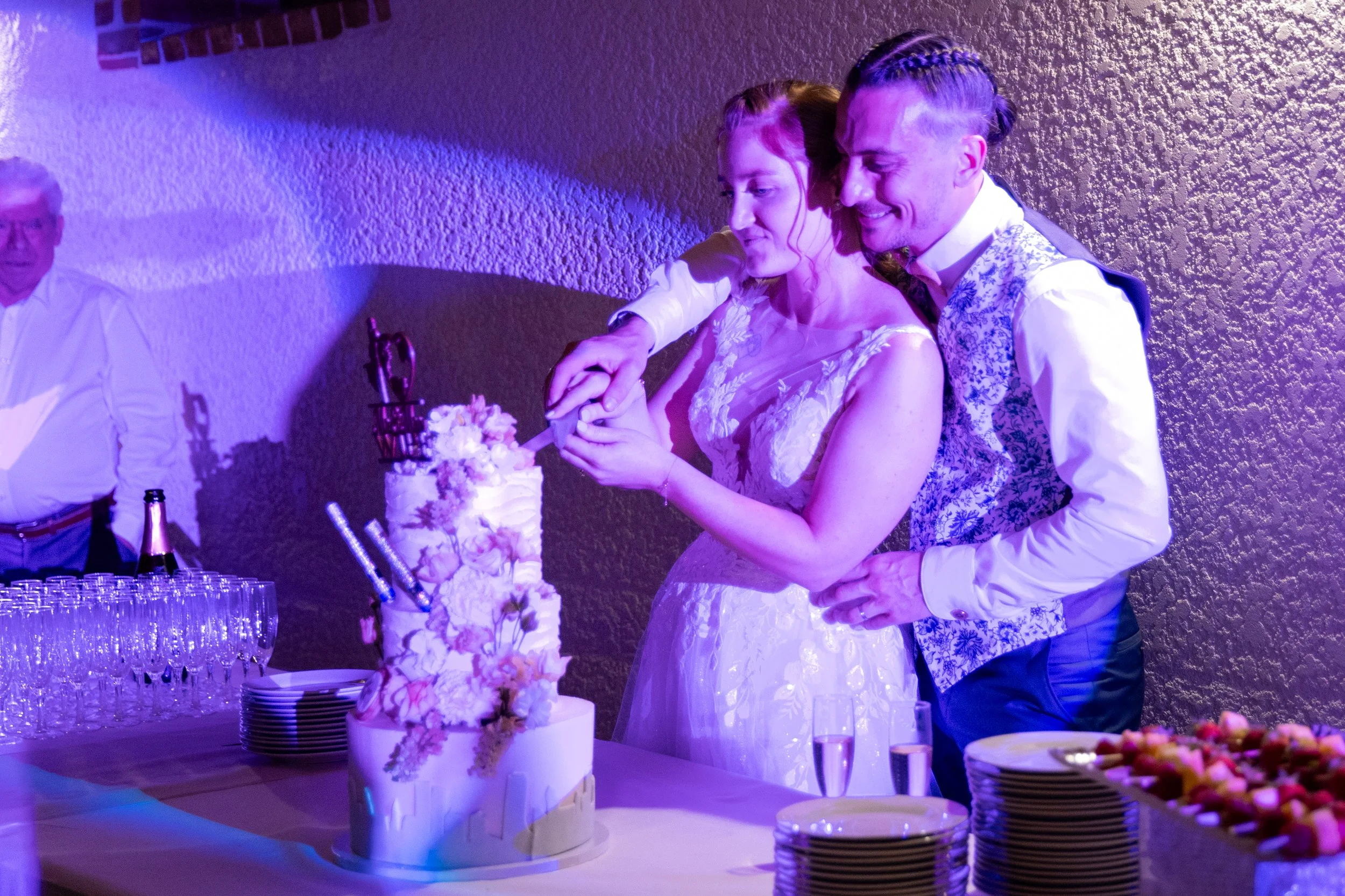 Un couple de mariés coupe leur gâteau de mariage sous des lumières violettes, avec des invités en arrière-plan.