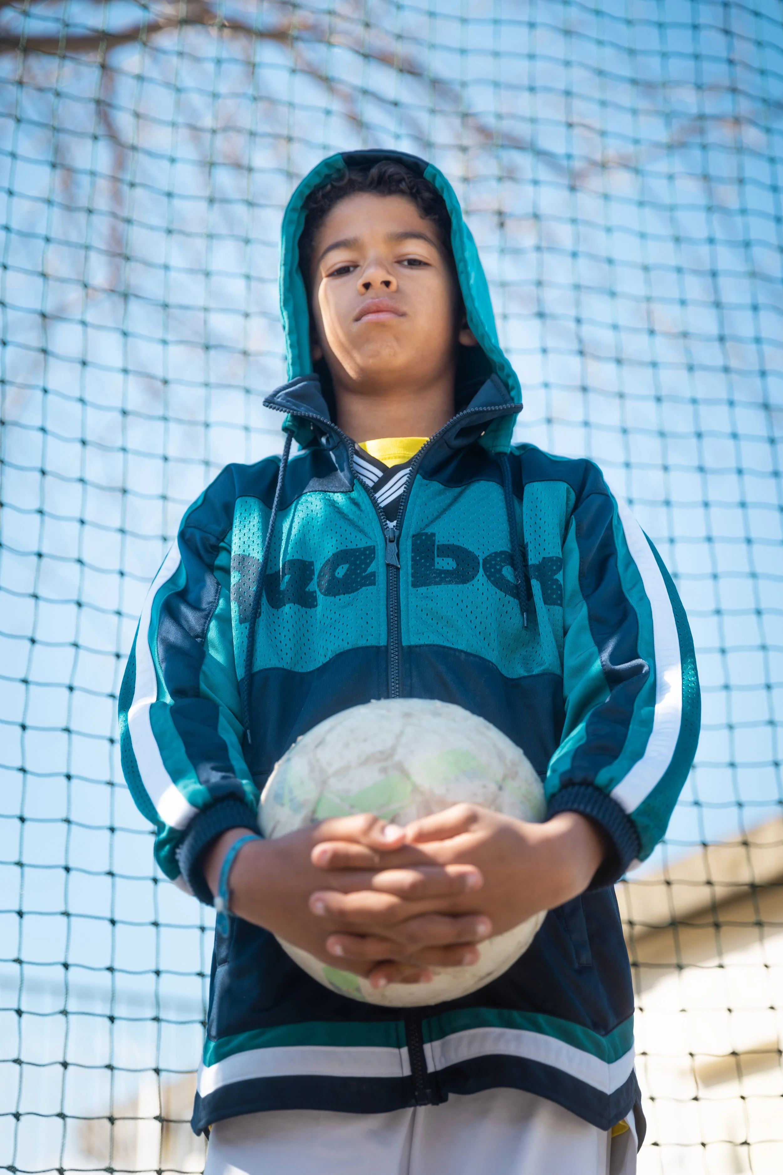Un garçon tenant un ballon de football, portant une veste à capuche bleue, debout devant un filet de but de football.