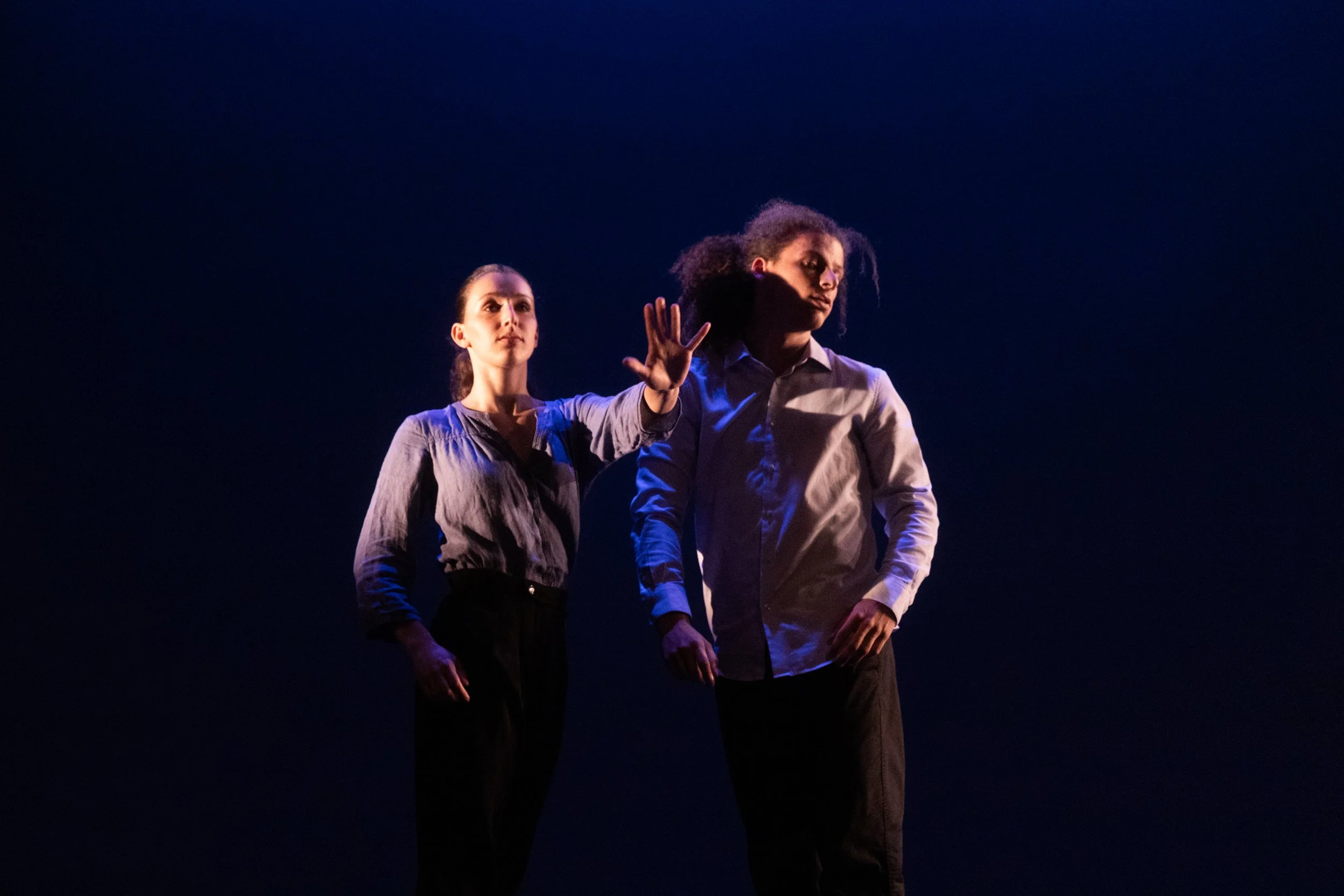 Deux personnes en costume sur scène lors d'une performance ou d'une danse, avec un fond sombre et un éclairage ciblé sur elles.