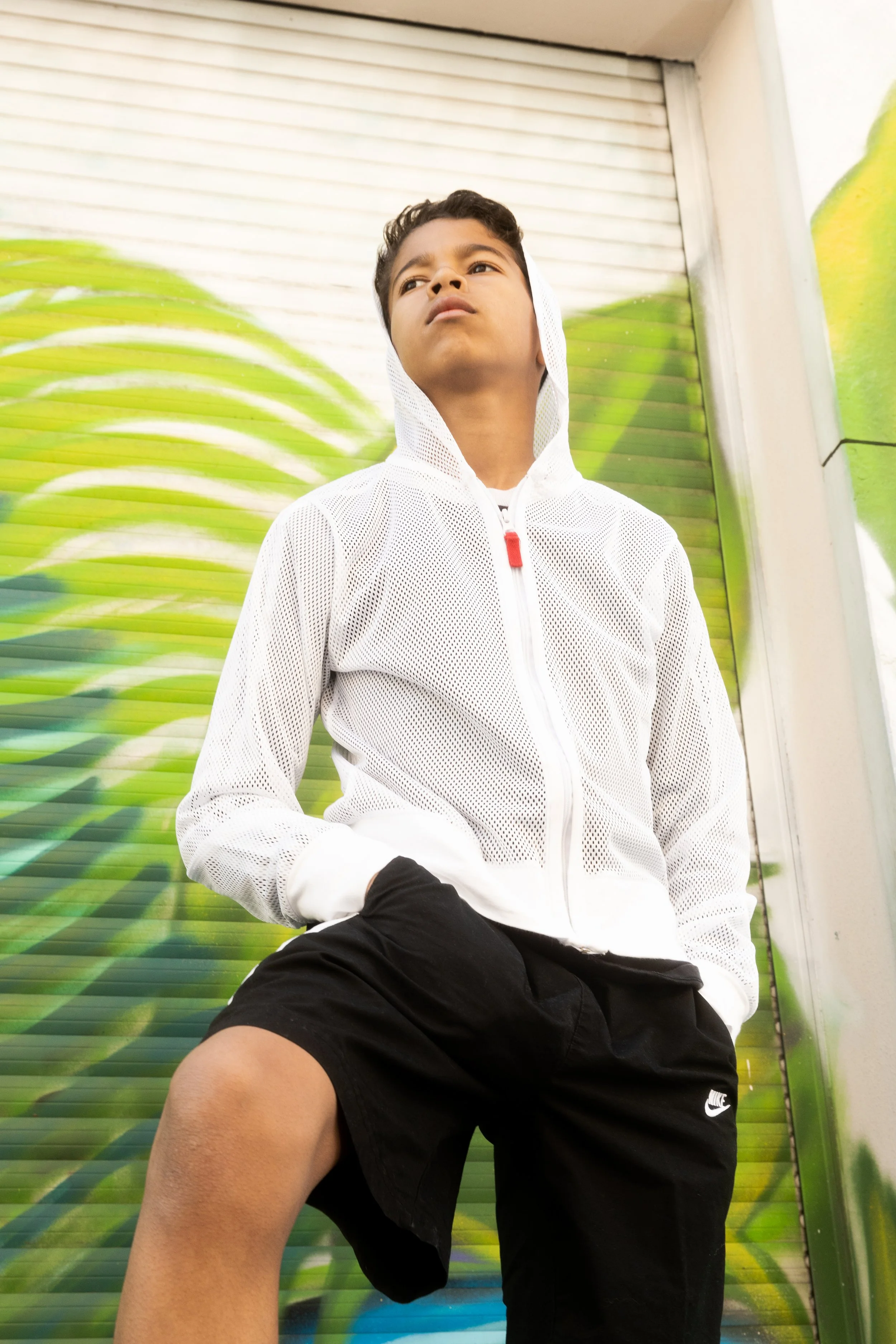 Jeune garçon portant un survêtement blanc à capuche et un short noir, posé contre un mur avec un graffiti de feuilles vertes.