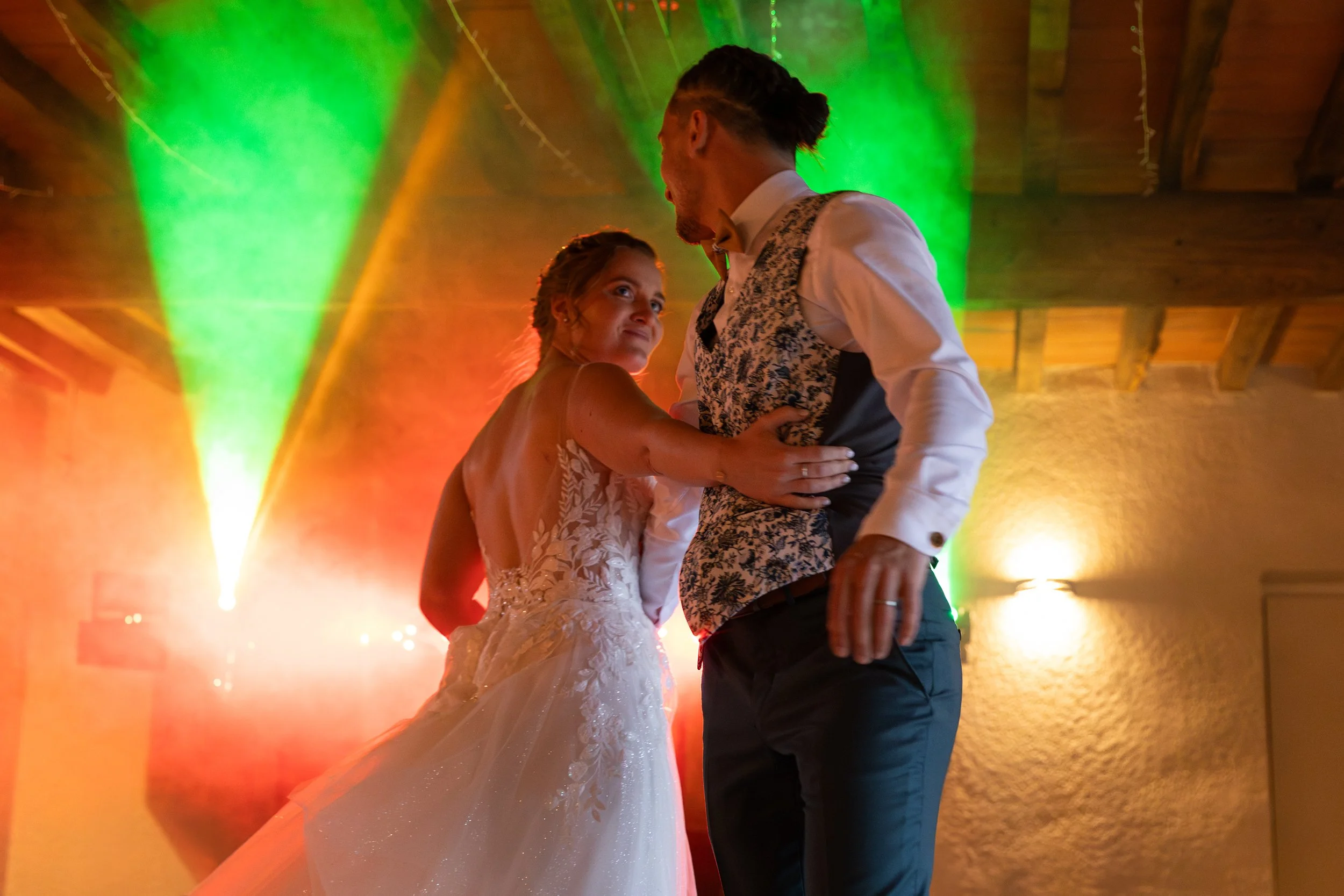 Un jeune couple danse lors d'un mariage, la femme porte une robe de mariée blanche et l'homme un veston à motif floral, avec des lumières colorées vertes, jaunes et rouges en arrière-plan.
