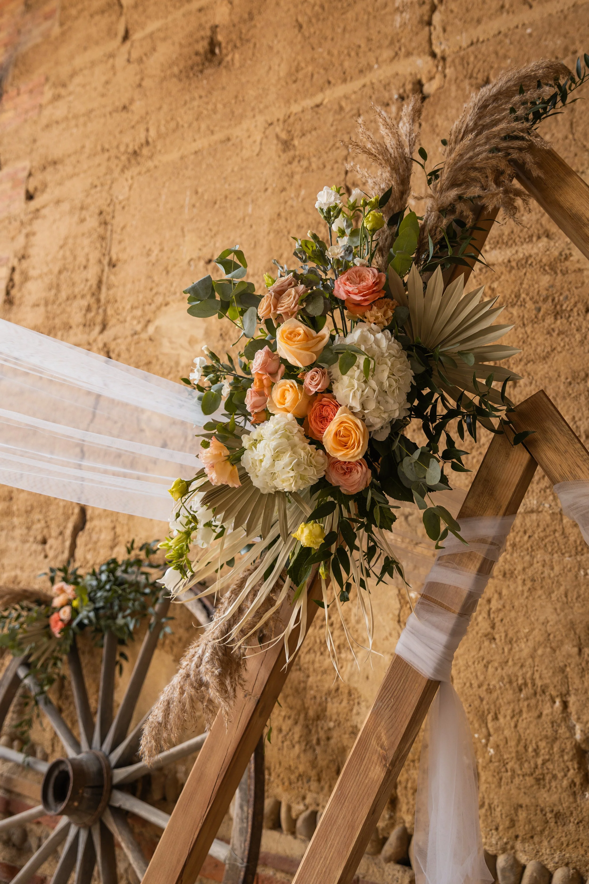 Un arrangement floral de mariage avec des roses, hortensias et feuillage, fixé à un escalier en bois avec des rubans de tissu blanc, contre un mur en pierre marron.