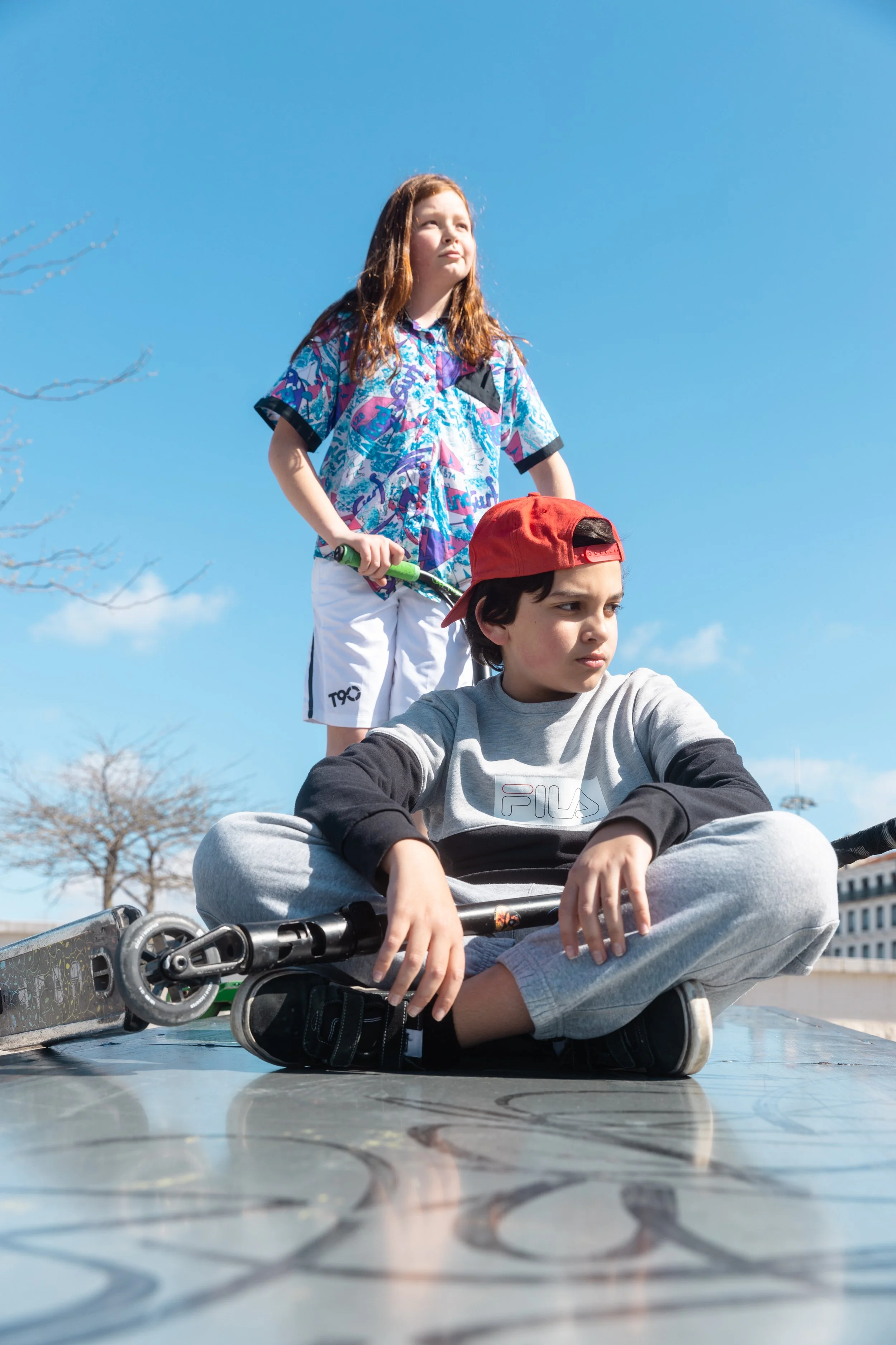 Deux enfants, un garçon assis et une fille debout, dans un parc lors d'une journée ensoleillée. La fille tient une patinette et regarde au loin, tandis que le garçon est assis sur le sol avec une expression pensive.