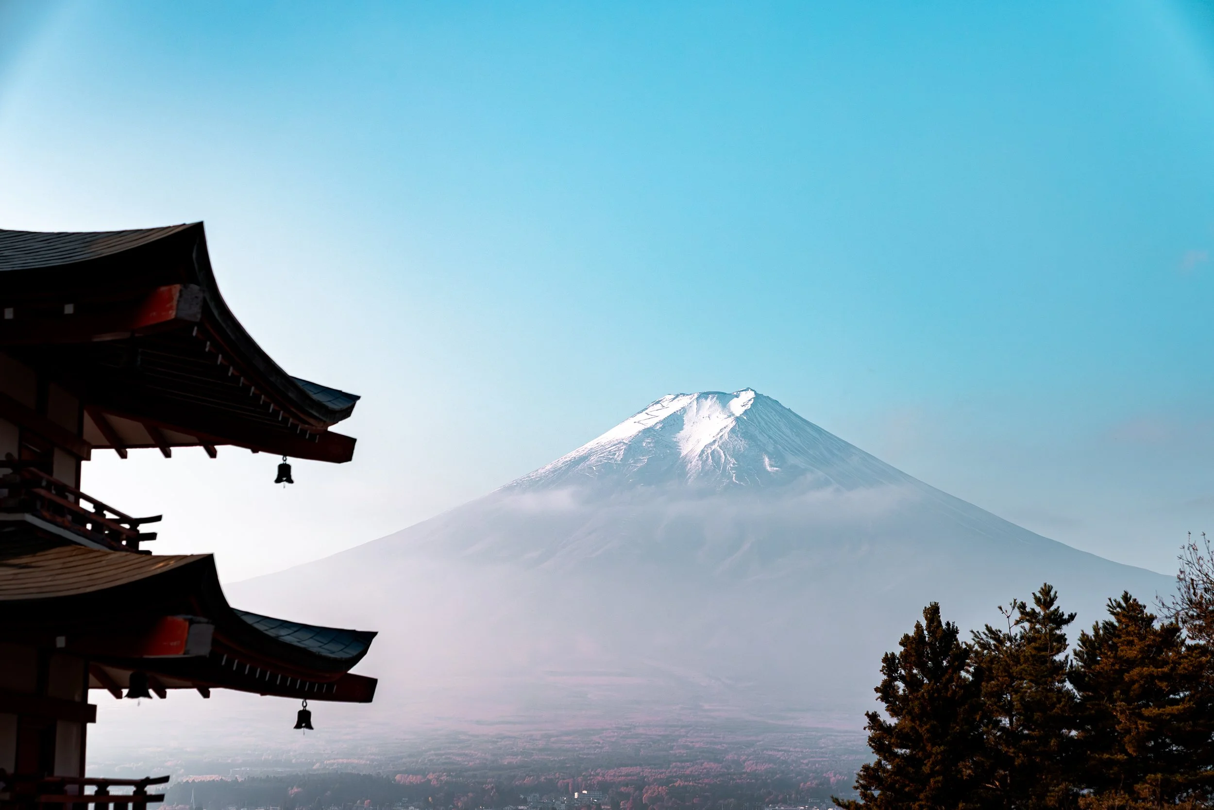 Mont Fuji enneigé visible en arrière-plan, avec un temple traditionnel japonais en premier plan à gauche et des arbres au premier plan en bas à droite.