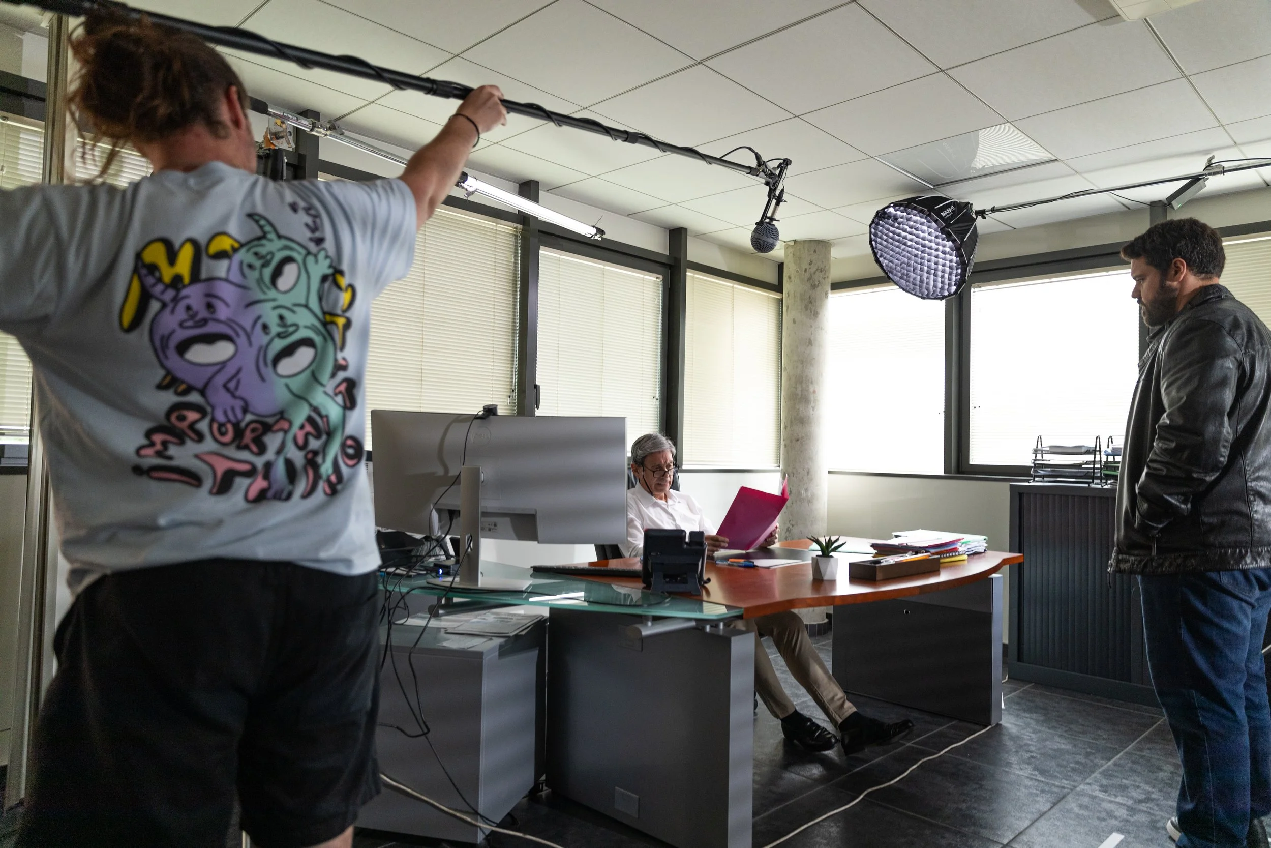 Une scène de tournage en intérieur avec un homme assis à un bureau, deux hommes debout et un équipement de tournage.