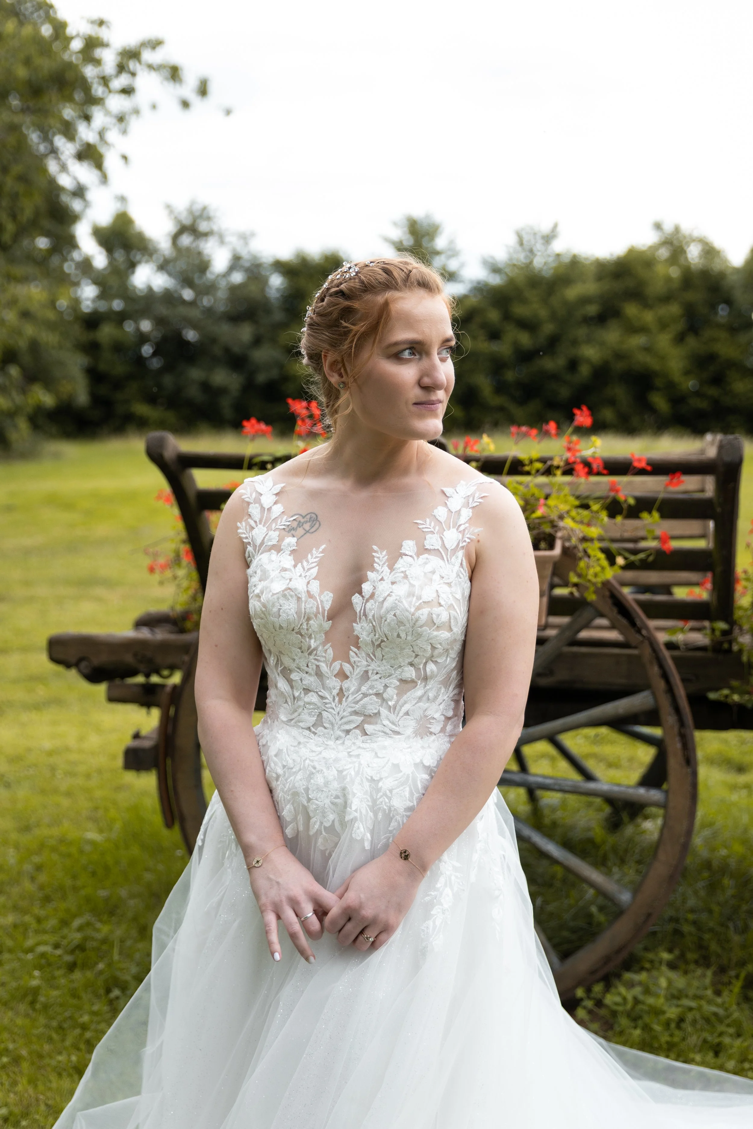 Une mariée en robe blanche avec des broderies florales, posant devant une brouette en bois avec des fleurs rouges, dans un jardin.