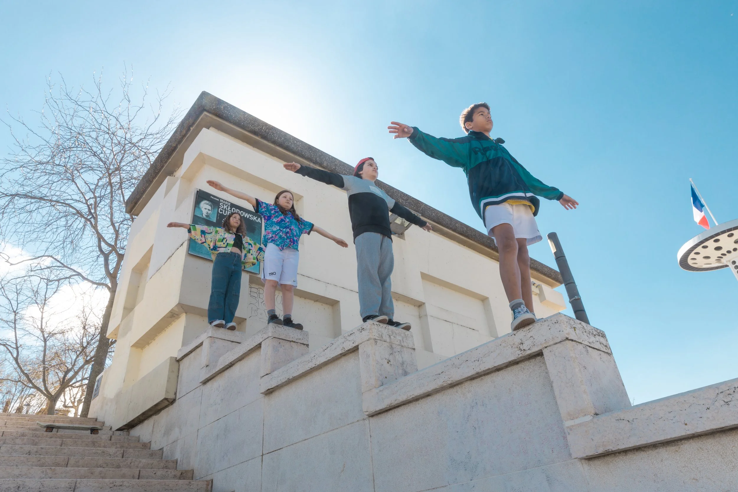 Quatre enfants debout sur des blocs de pierre en position de vol avec les bras étendus, face à un bâtiment blanc, sous un ciel bleu clair, avec des arbres sans feuilles en arrière-plan.