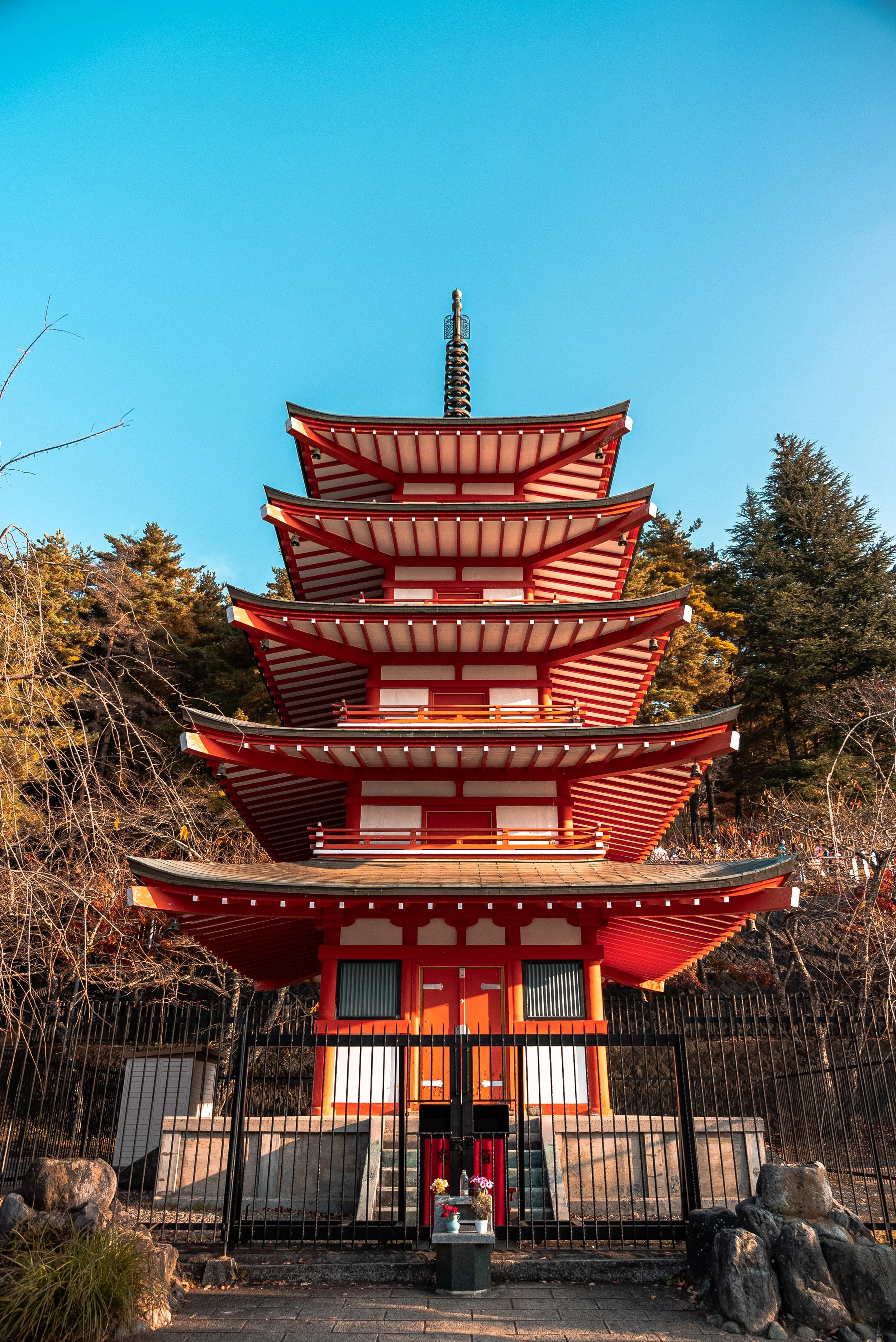 Pagode japonaise rouge avec cinq étages, entourée d'une clôture en fer noir et située devant un fond de forêt, sous un ciel bleu clair.