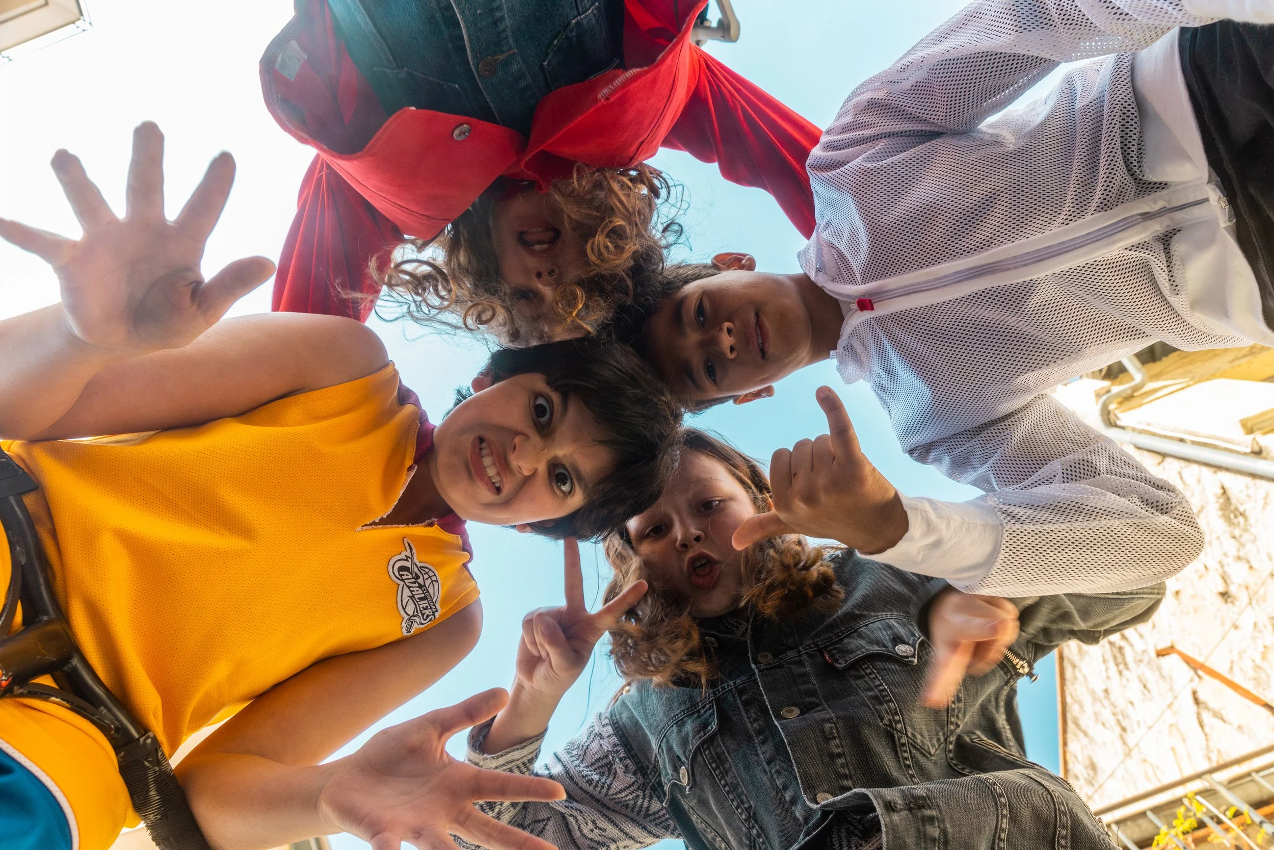 Groupe de quatre enfants regardant vers la caméra, prise du sol avec un ciel bleu en arrière-plan, expression joyeuse et gestes variés