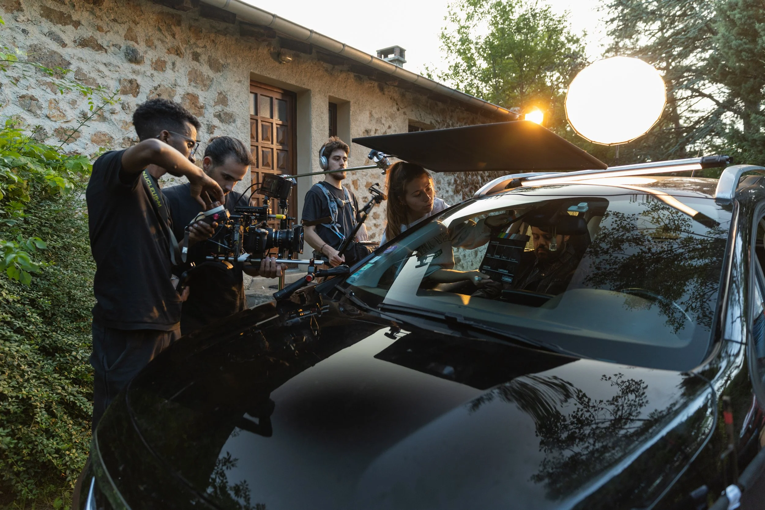 Une équipe de tournage en plein extérieur au coucher du soleil, utilisant une caméra professionnelle sur une voiture noire, avec une femme ajustant l'équipement et un homme dans la voiture en train de travailler sur un ordinateur portable, sous un gr