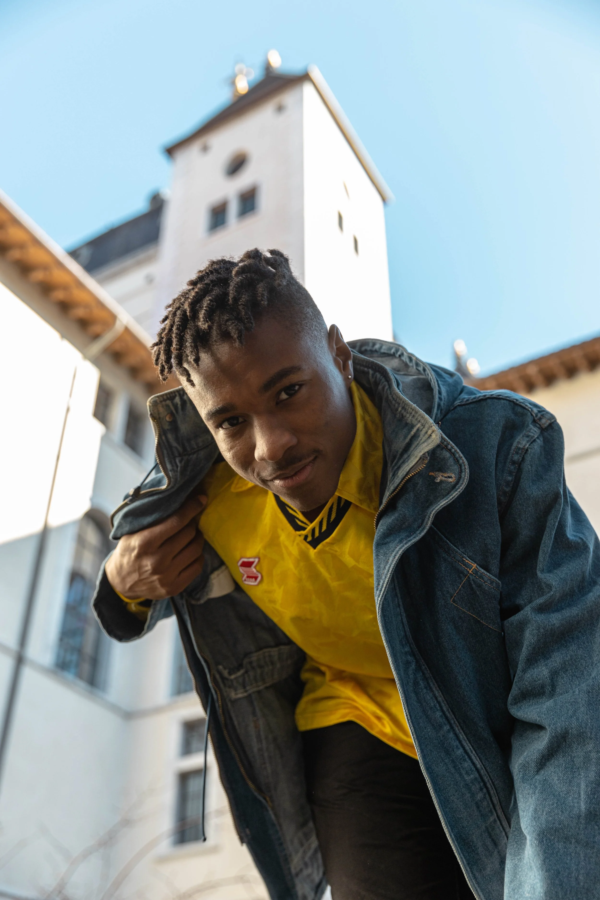 Jeune homme portant une veste en denim et un pull jaune, regardant l'objectif, avec un bâtiment en arrière-plan et un ciel bleu.