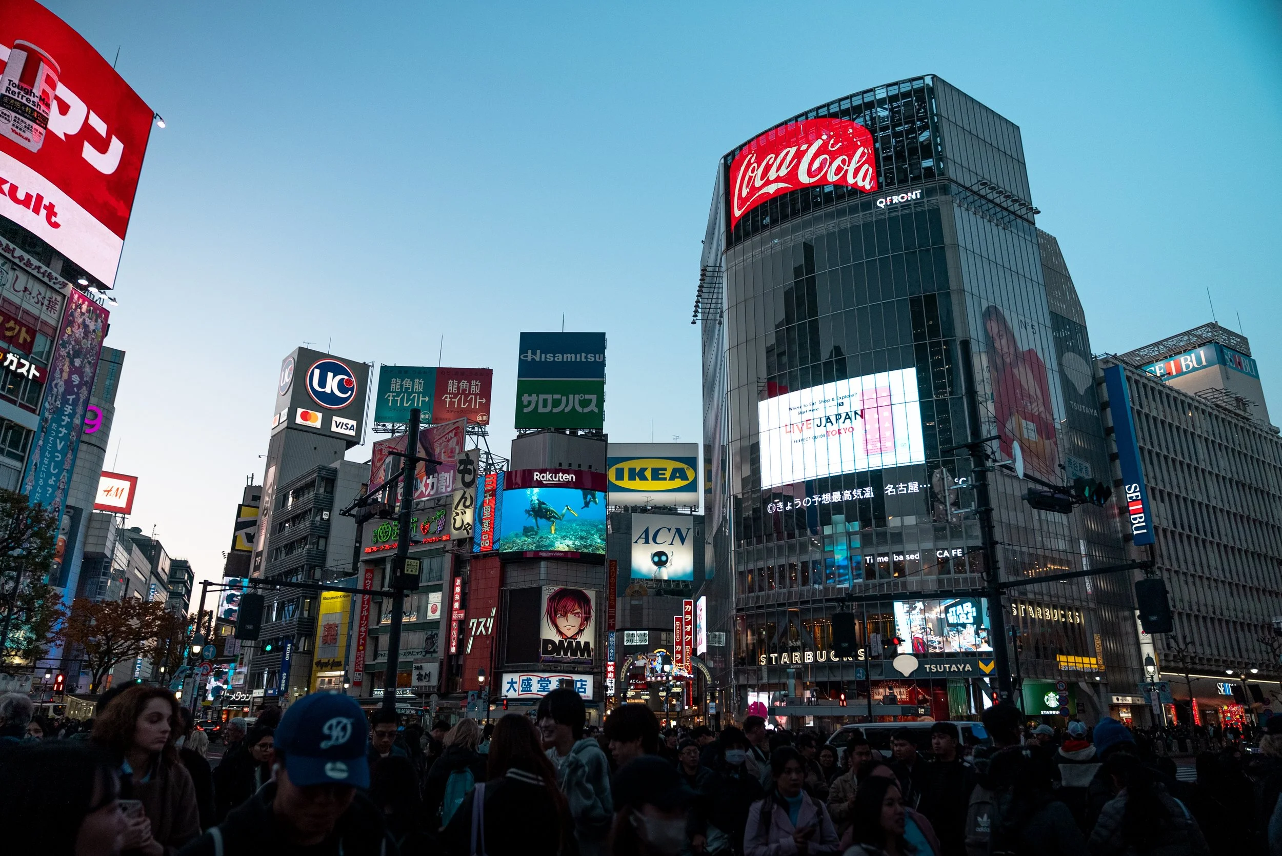 Vue panoramique d'une grande place urbaine avec de nombreux écrans publicitaires lumineux, notamment une grande affiche Coca-Cola, et une foule importante de personnes, dans un quartier animé de Tokyo, Japon.