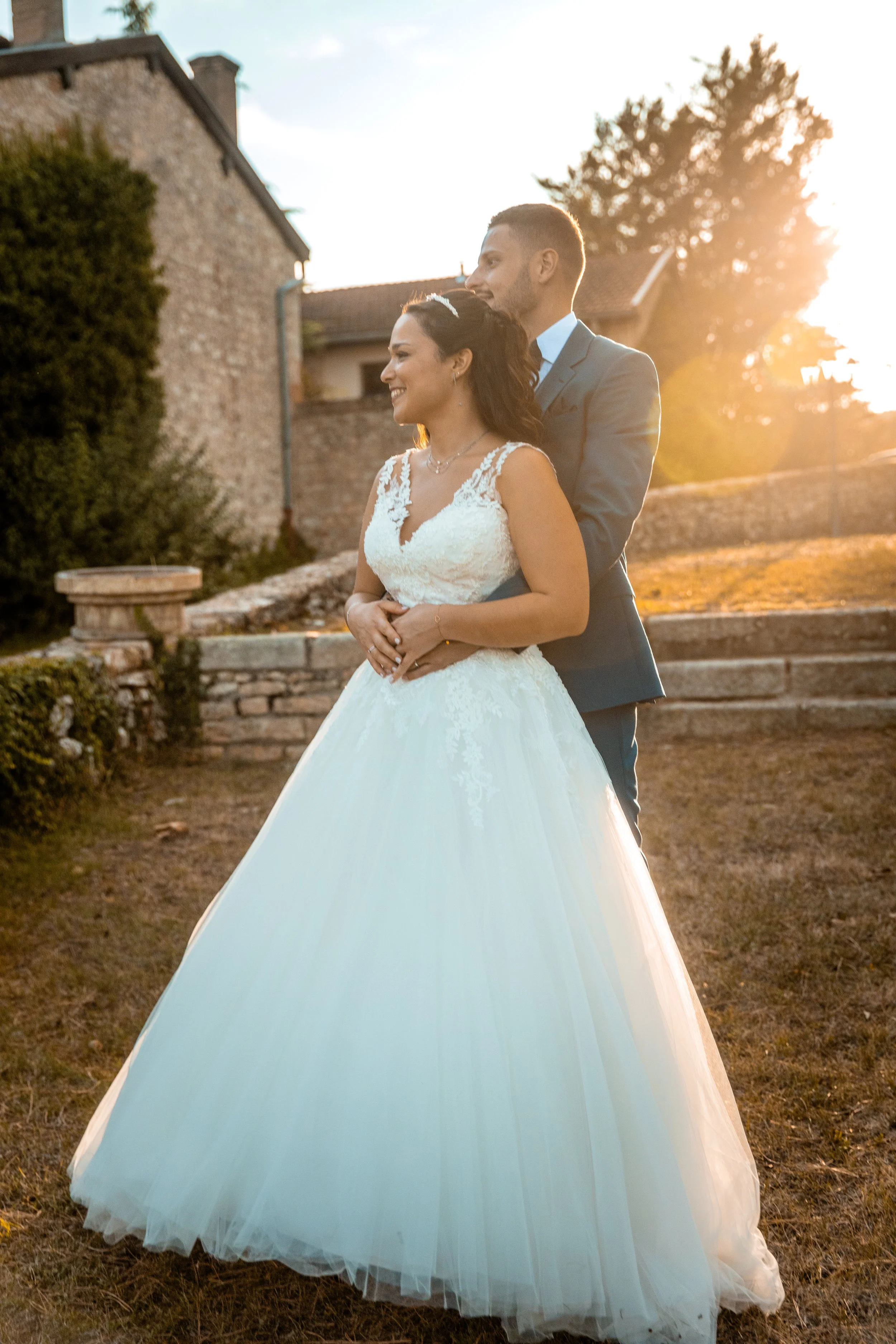 Un couple de mariés lors d'une séance photo en extérieur au coucher du soleil, avec une femme en robe de mariage blanche et un homme en costume gris, devant un vieux bâtiment en pierre et un arbre.