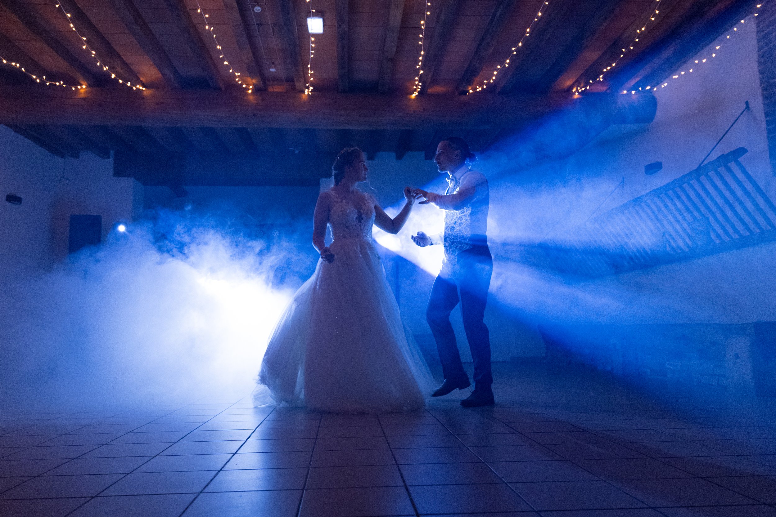 Un couple en habits de mariage danse sous des lumières bleues et des guirlandes lumineuses dans une salle de réception, avec un sol en carreaux.