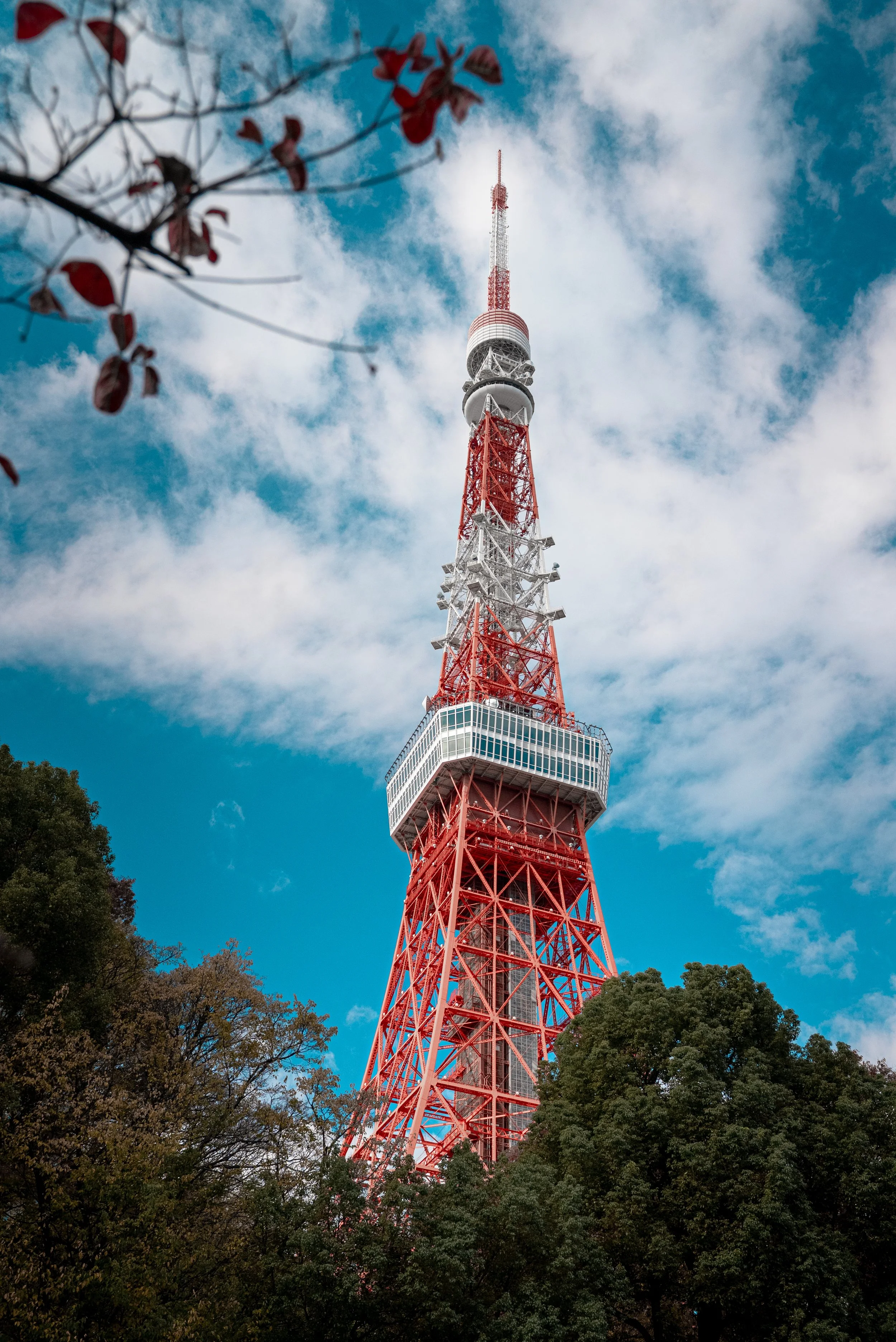 Une tour de transmission rouge et blanche se dresse contre un ciel bleu avec des nuages, entourée d'arbres verts.