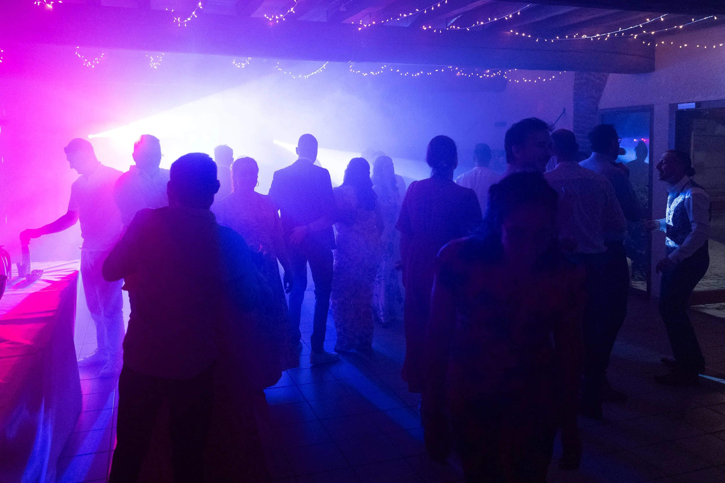Groupe de personnes dansant dans une fête avec éclairage coloré et guirlandes lumineuses au plafond.