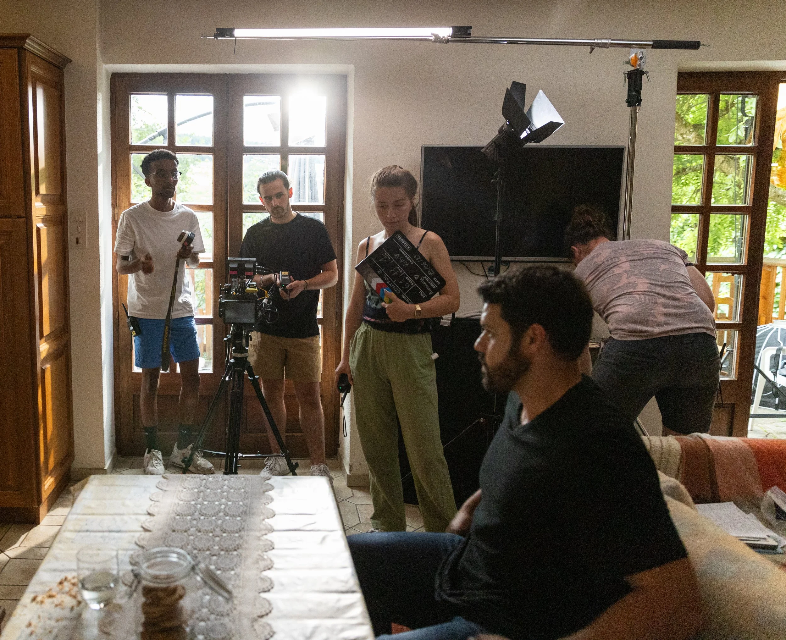 Une équipe de tournage en intérieur, avec des caméras et des éclairages, préparant une prise de vue près d'une porte-fenêtre, avec trois personnes en train de se préparer ou de superviser le tournage.