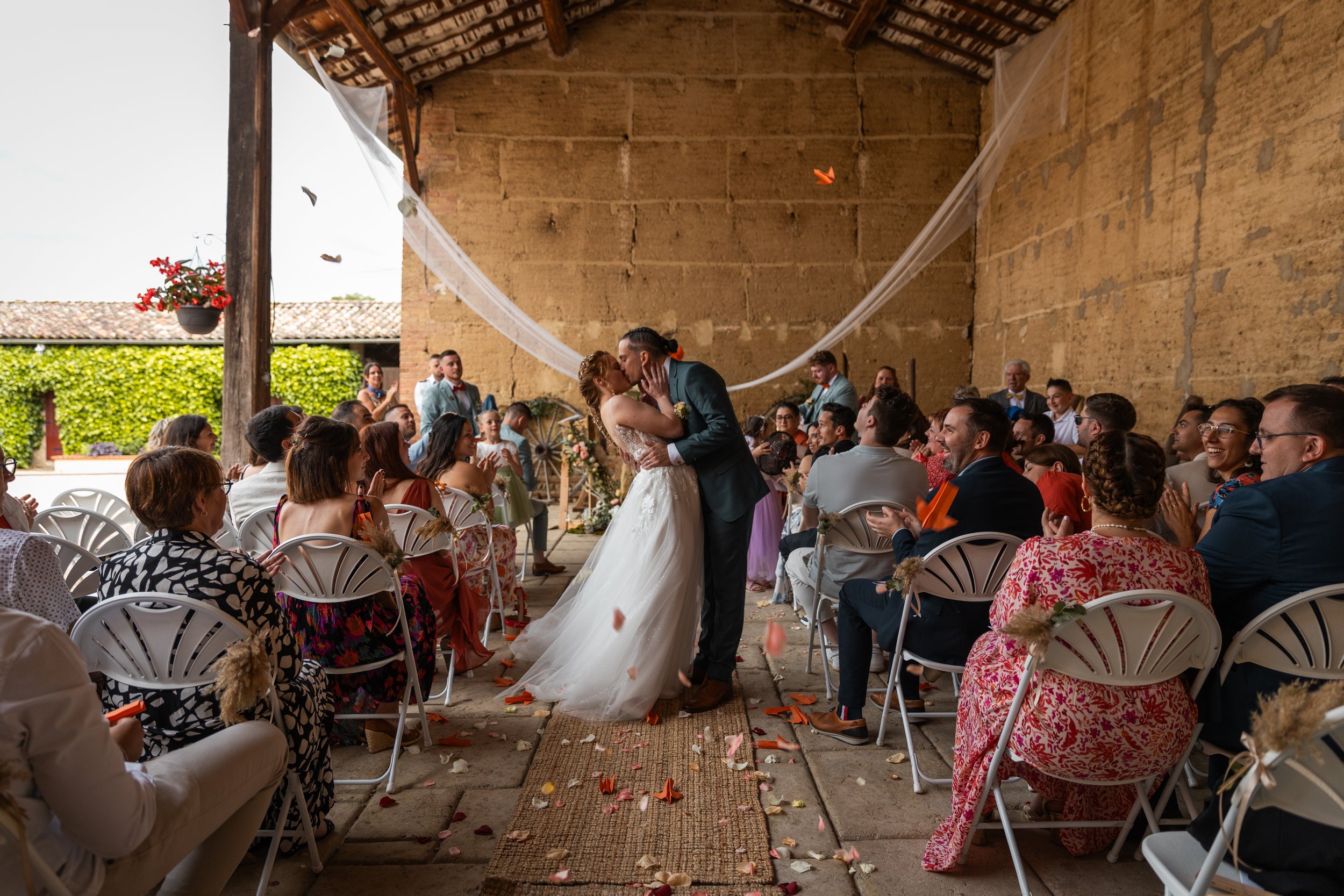 Les mariés s'embrassent lors de leur cérémonie de mariage dans un espace en plein air avec des invités assis, décorations de fleurs et toile blanche en arrière-plan.