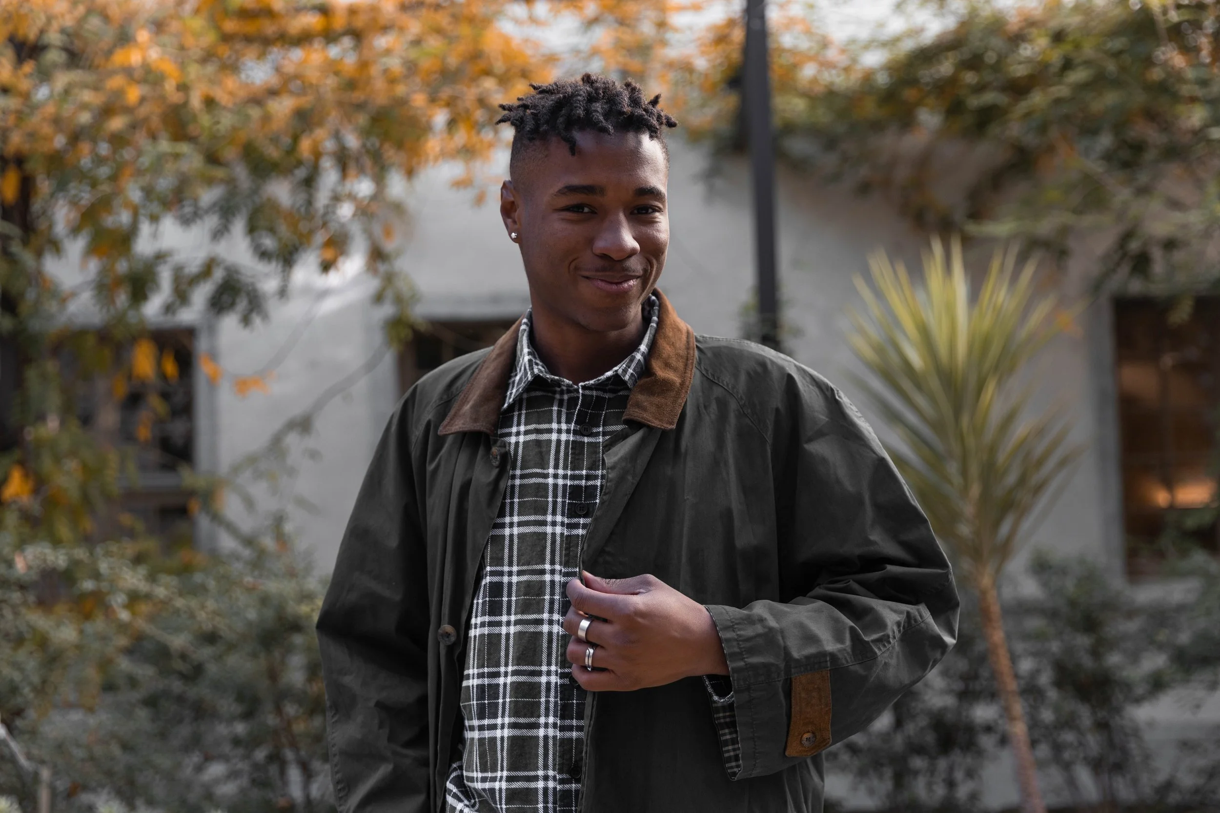 Jeune homme souriant portant un manteau noir avec col en cuir marron, une chemise à carreaux noire et blanche, et des boucles d'oreilles dans un jardin avec des arbres aux feuilles orange.