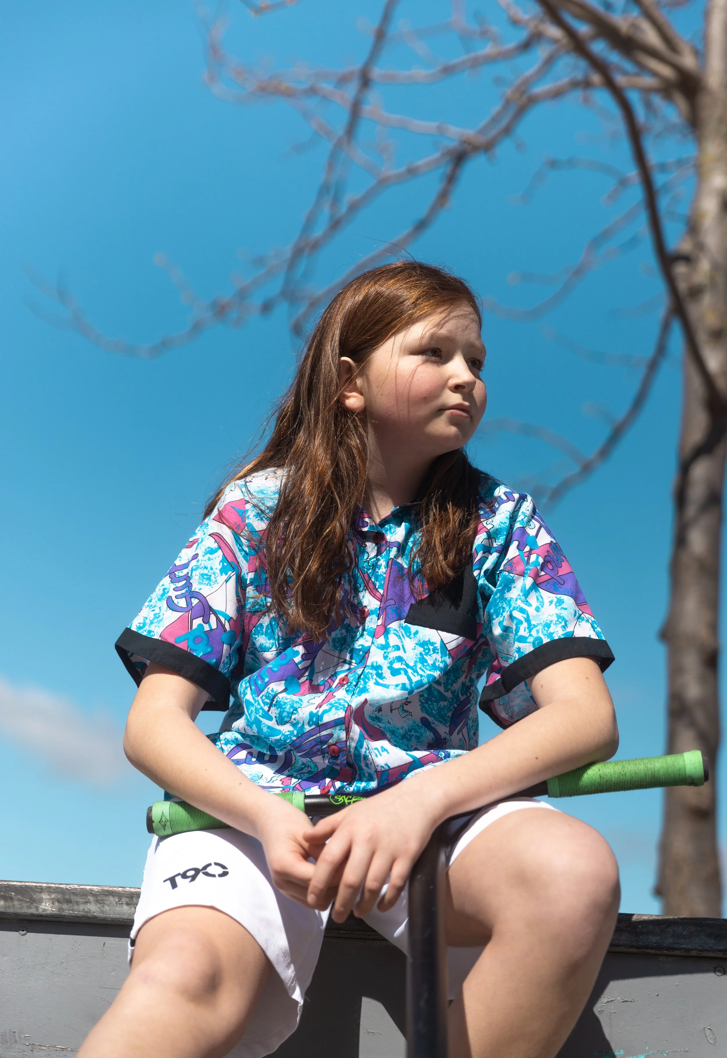 Jeune fille aux cheveux longs assise dans une barque, portant un maillot coloré et un short blanc, sous un ciel bleu avec des branches d'arbre.