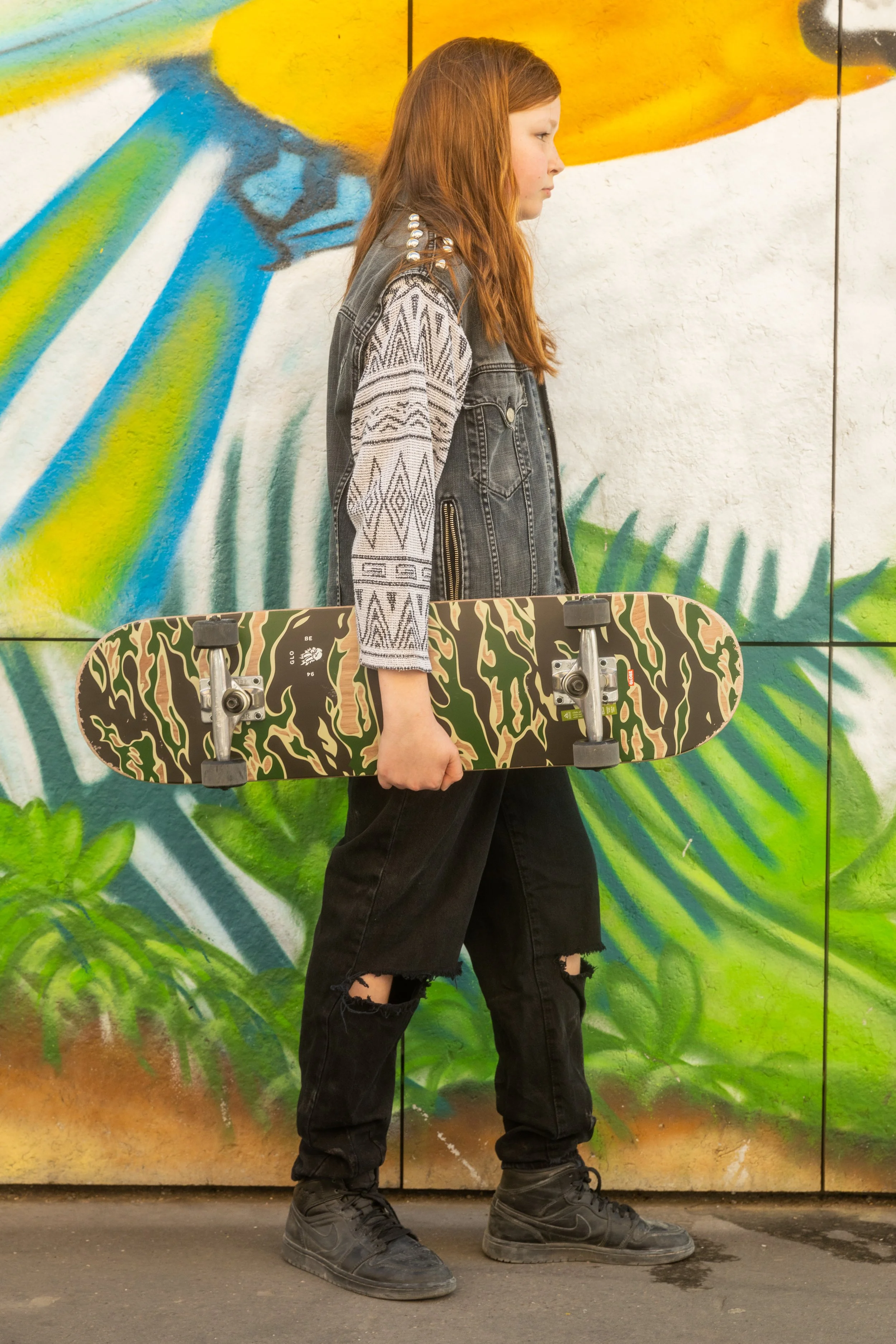 Une jeune fille aux cheveux roux, portant un gilet en jean avec des manches à motif, des pantalons noirs déchirés aux genoux et des bottes noires, tient une planche de skateboard à la main, devant un mur peint avec un graffiti coloré.