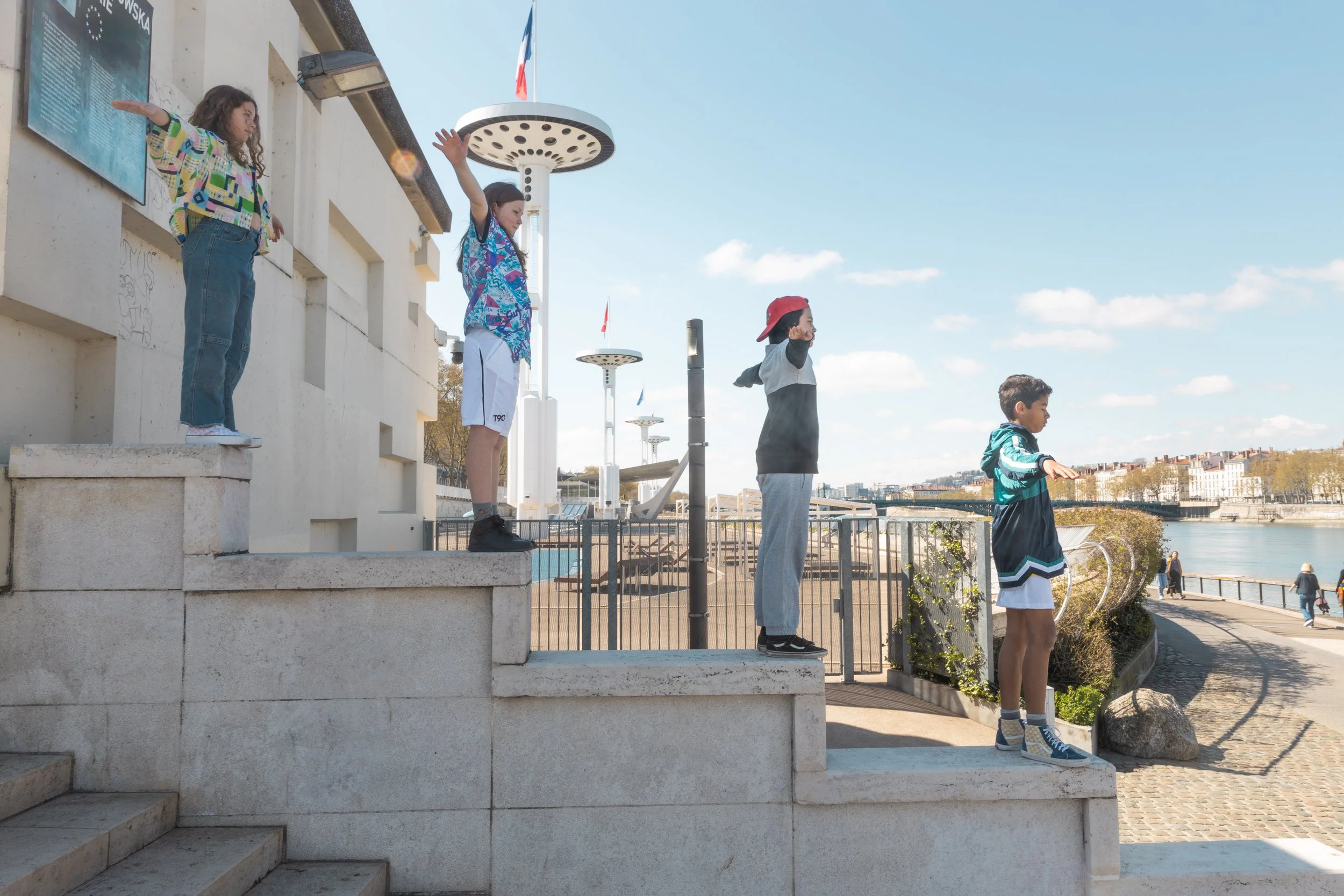 Cinq enfants jouent sur des marches près d'une rivière en plein air, avec un ciel bleu et quelques nuages. Ils font des gestes de salut ou de bras levés, et il y a des lampadaires modernes et un drapeau français en arrière-plan.