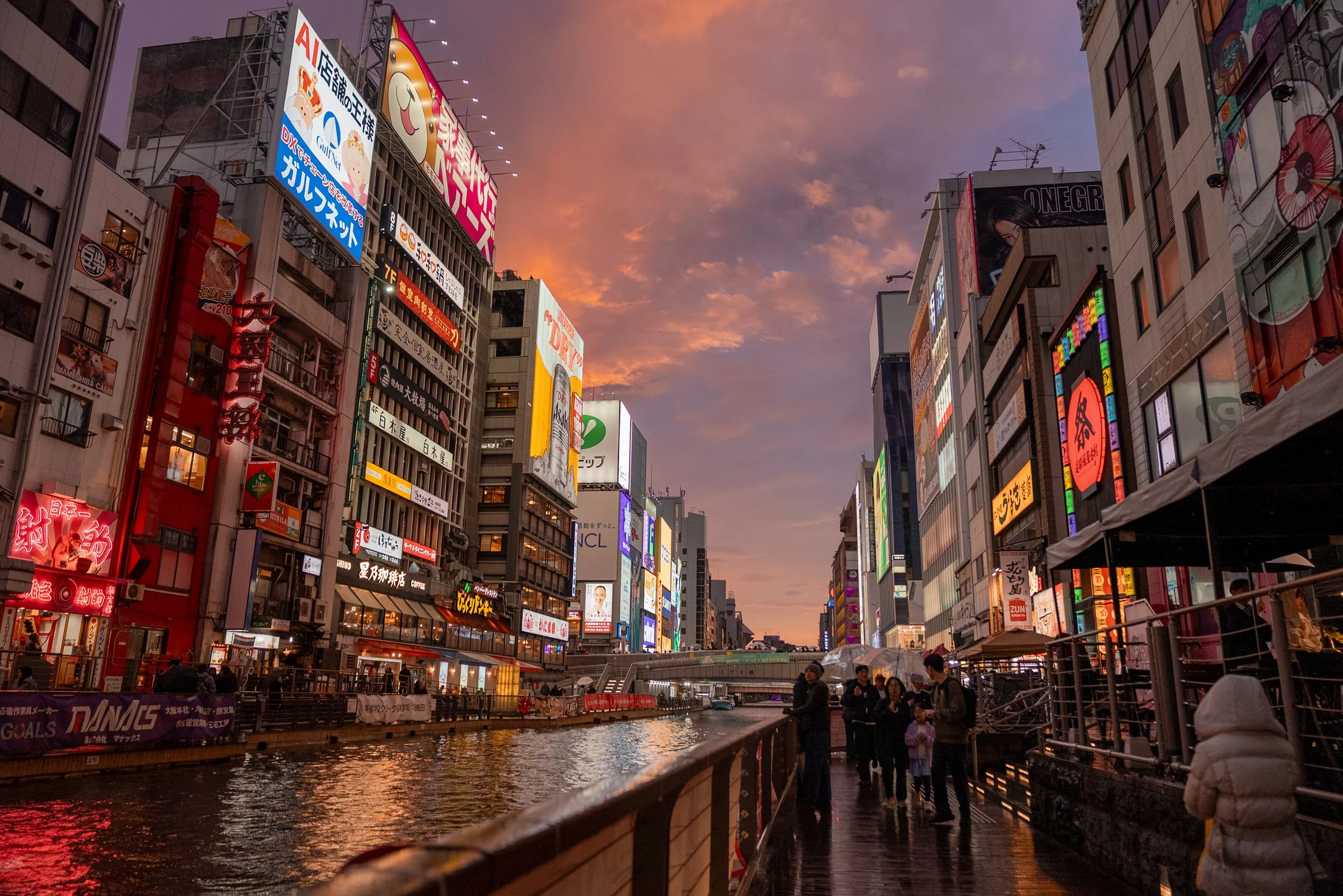 Vue d'une rue animée avec des gratte-ciel surplombant la rivière à l'heure du coucher du soleil, illuminée par des panneaux publicitaires colorés en japonais
