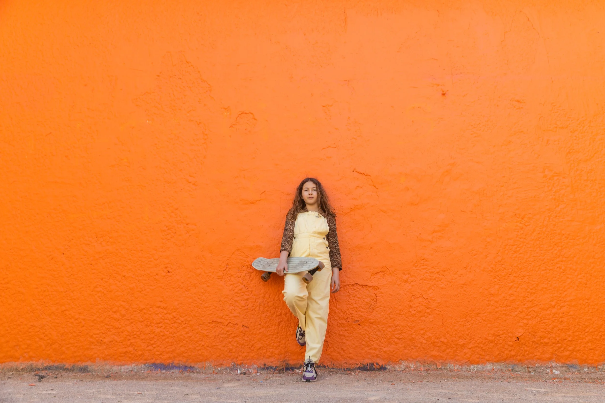 Jeune fille debout contre un mur orange, tenant un skateboard.