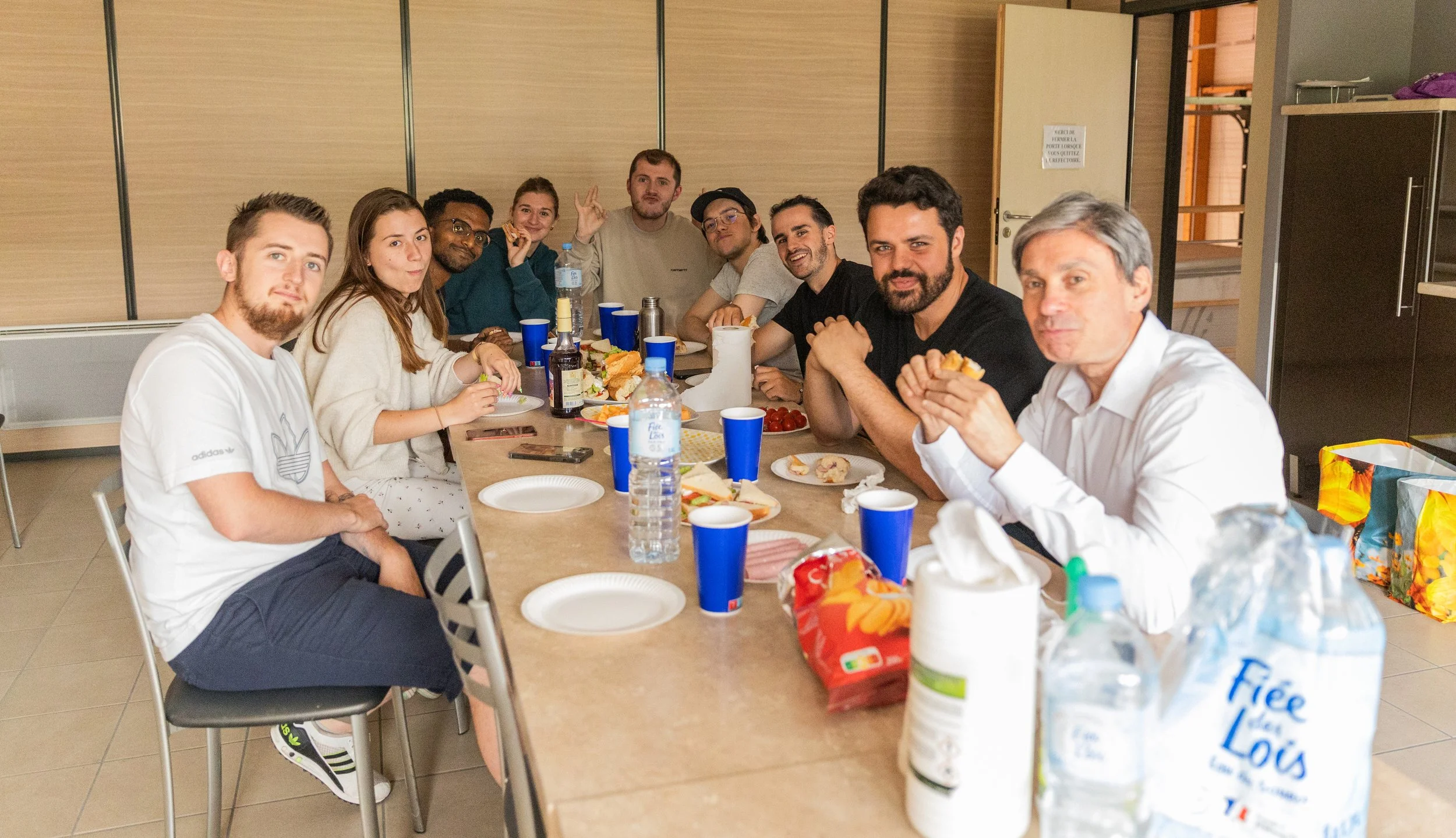 Groupe de personnes autour d'une table lors d'une pause déjeuner, en train de manger et de discuter, dans une salle intérieure.