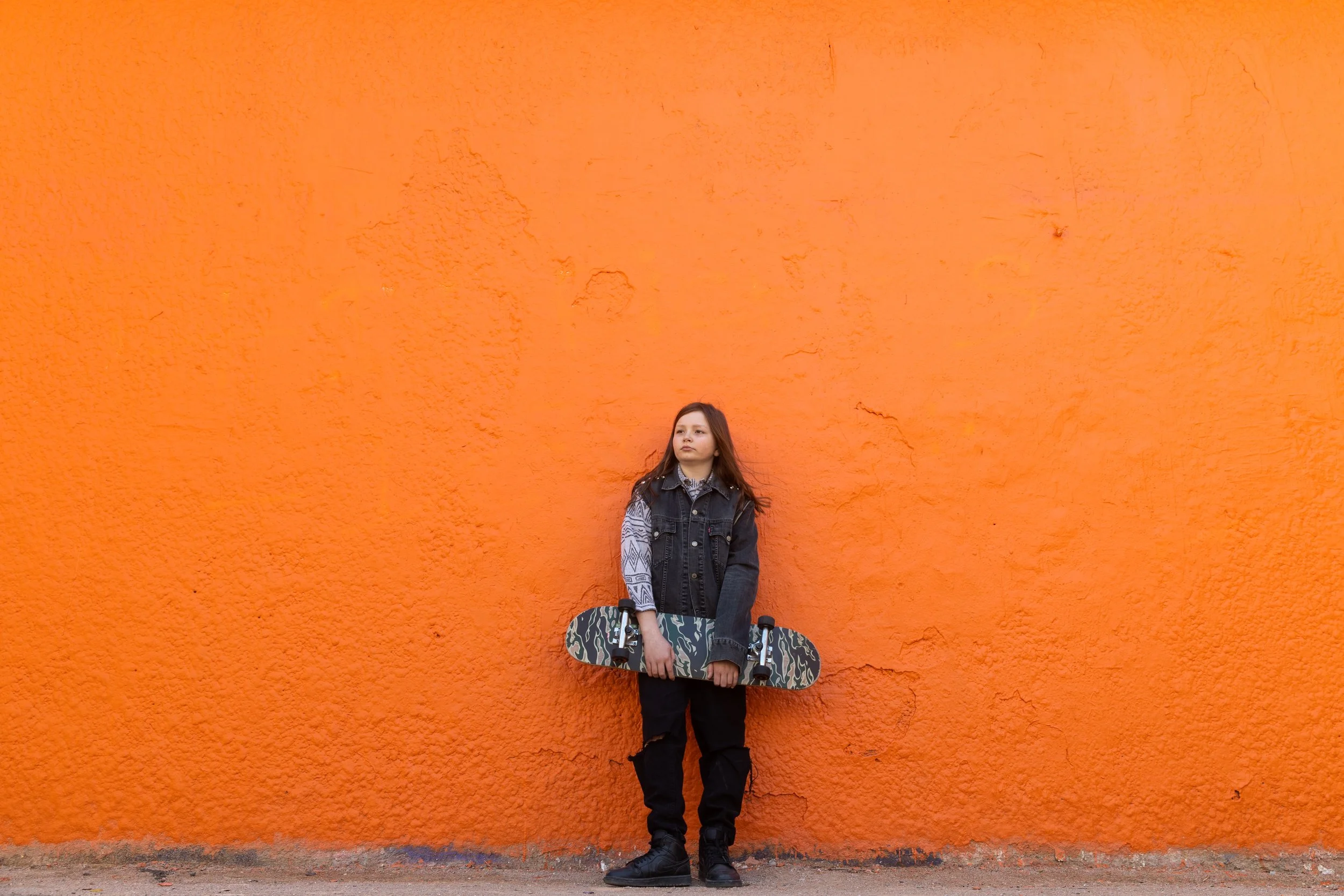 Jeune fille tenant une planche à roulettes devant un mur orange.