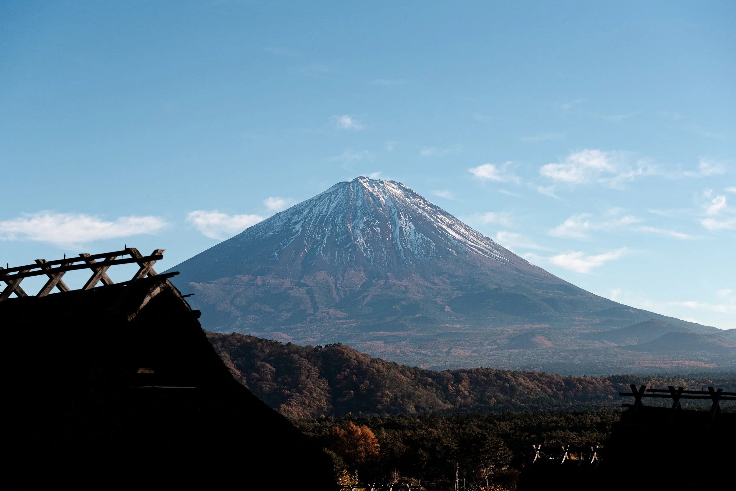 Vue du mont Fuji avec du ciel bleu et quelques nuages, en arrière-plan, avec des toits en bois en silhouette au premier plan.