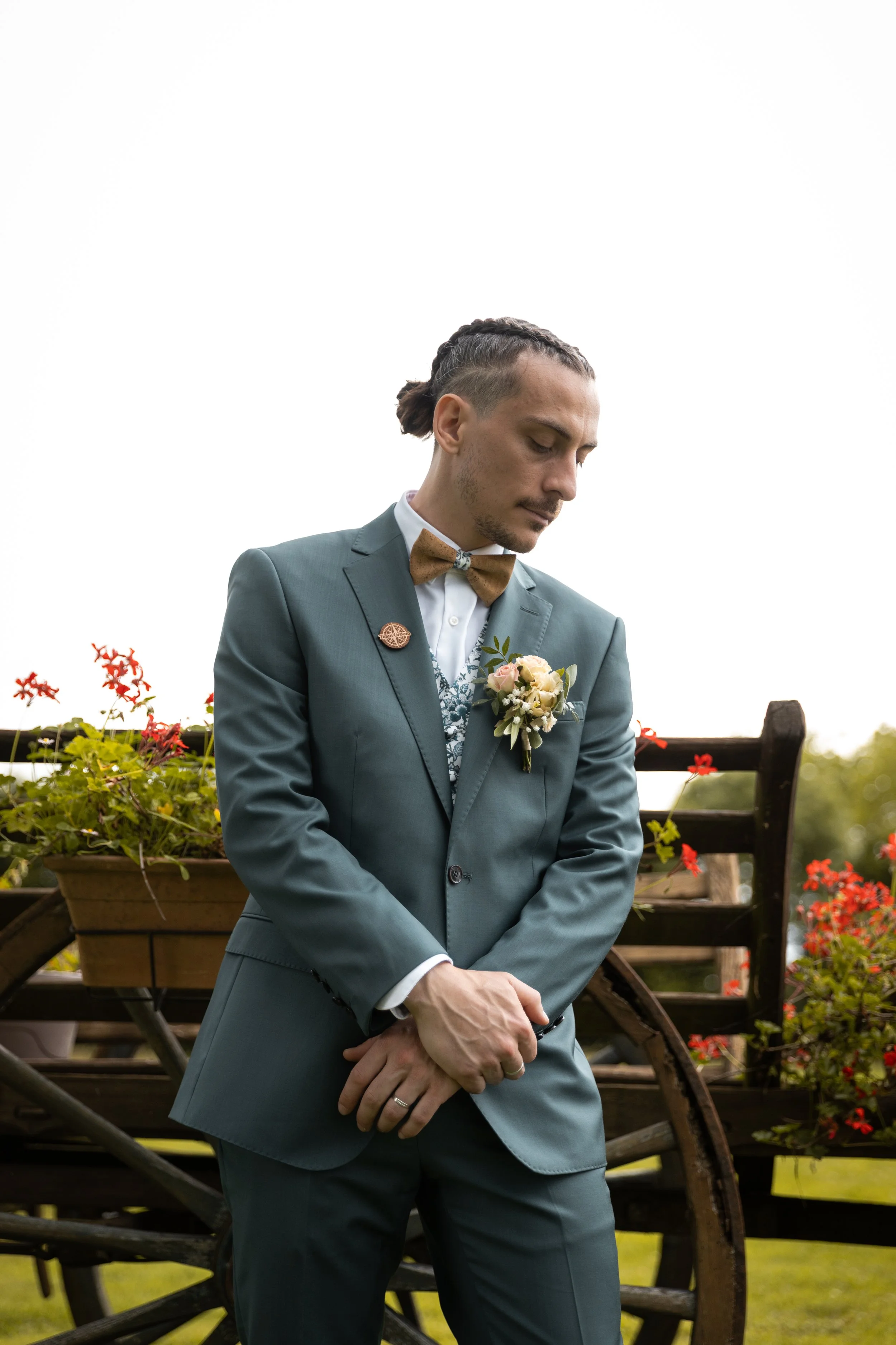 Un homme en costume gris avec un nœud papillon marron et une petite boutonnière, debout devant un vieux chariot en bois avec des fleurs rouges et jaunes. Il semble pensif, avec ses mains croisées et la tête inclinée vers le bas.