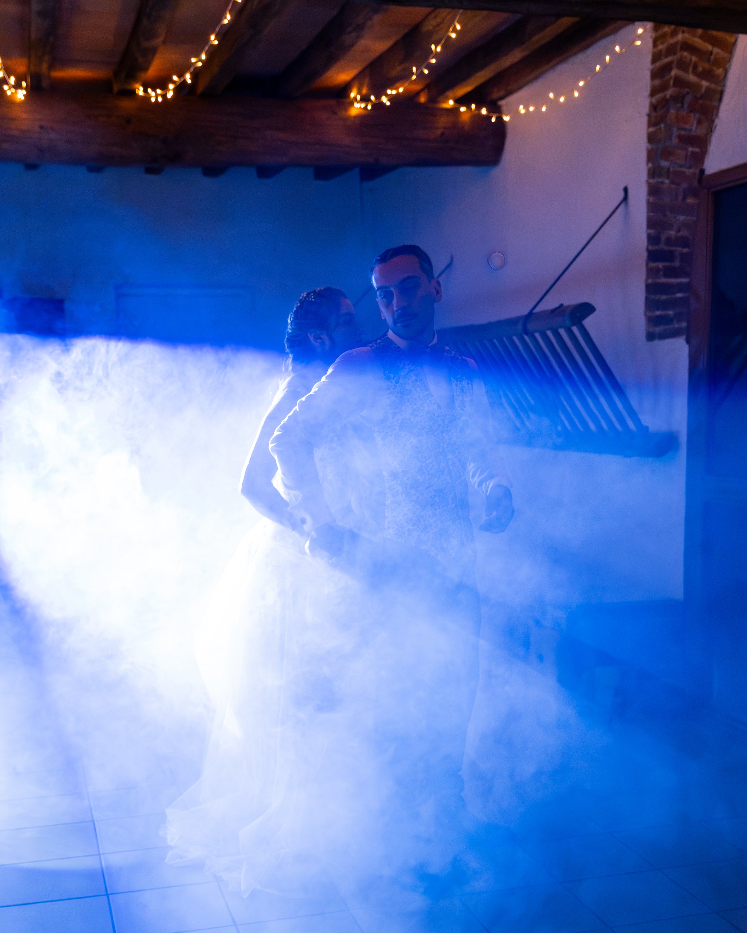 Un couple danse dans une pièce sombre avec un éclairage bleu et des guirlandes lumineuses au plafond, entourés de brouillard ou de fumée.