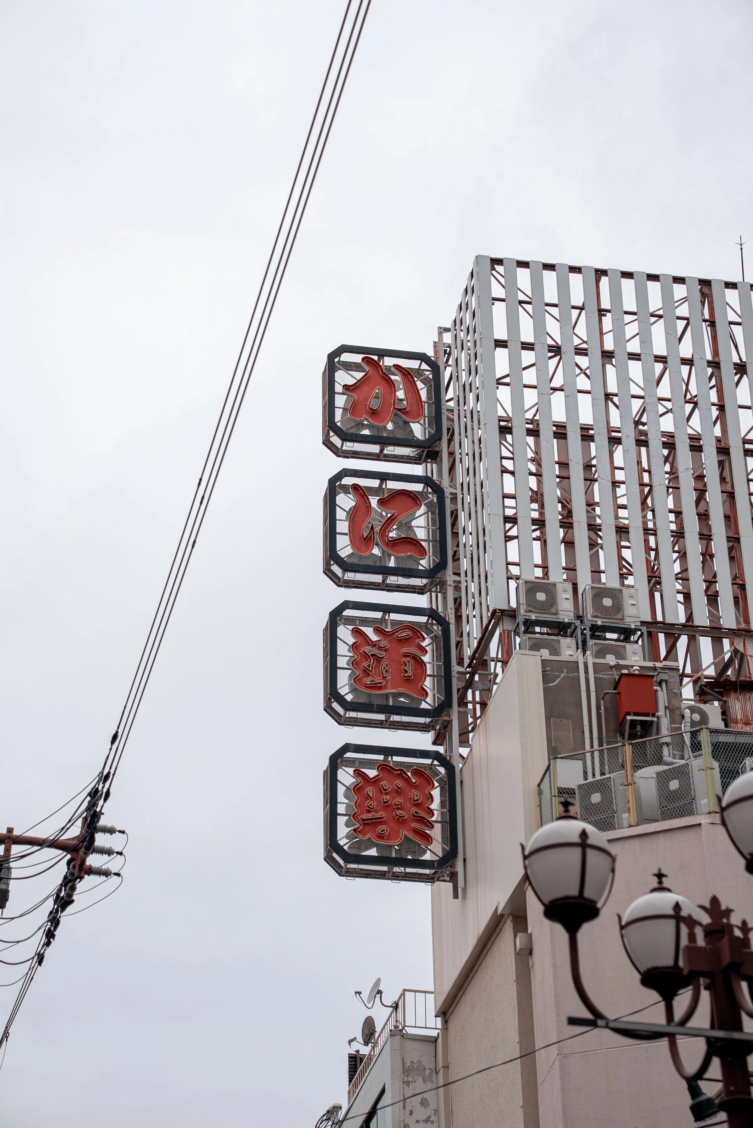Un panneau en anglais avec des caractères japonais et chiffres en rouge, suspendu à un bâtiment urbain, avec un ciel gris et des lampadaires devant.