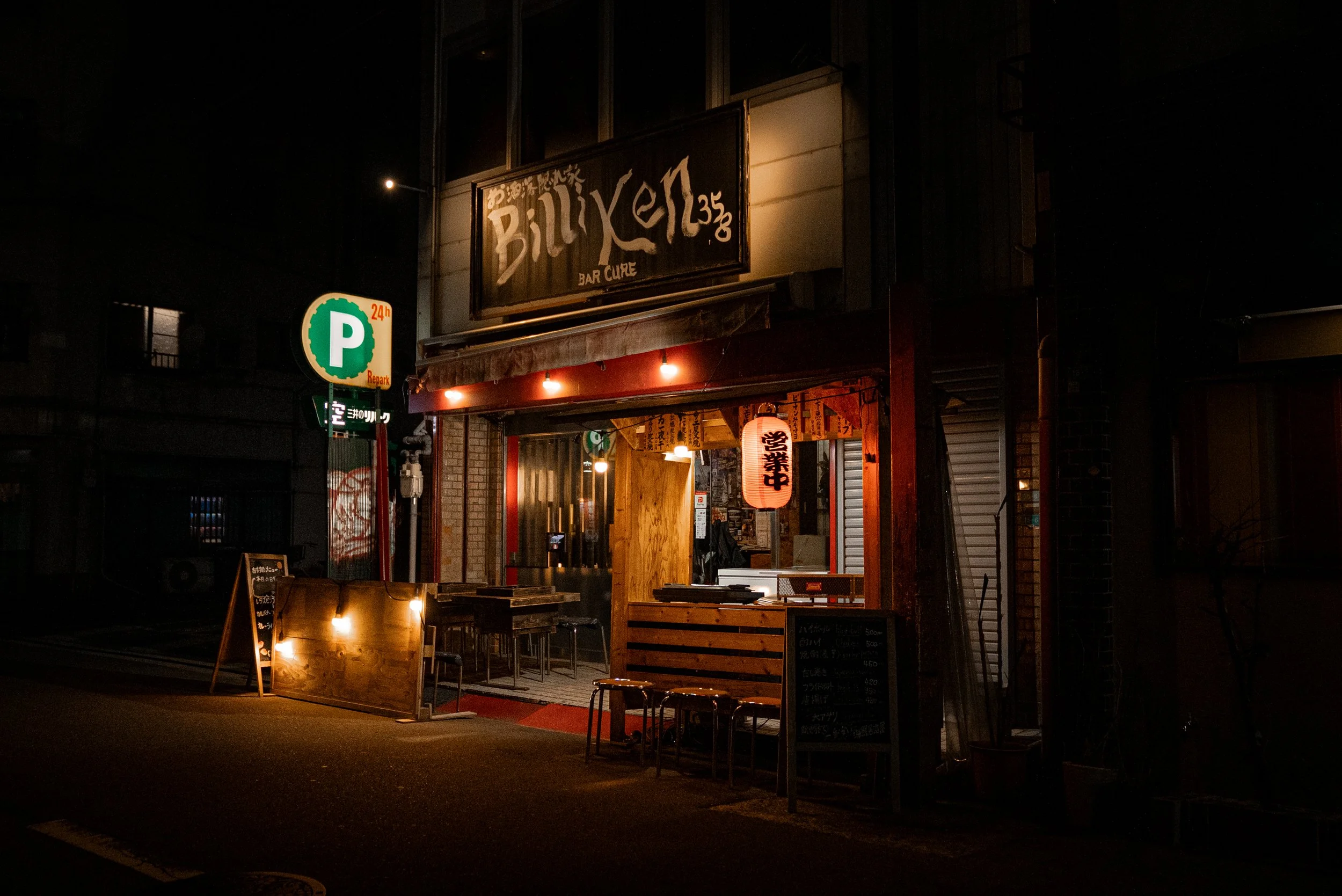 Façade d'un bar japonais appelé 'Billyken', éclairée par des lanternes et des lumières, avec un tableau de menu à l'extérieur et un panneau de parking.