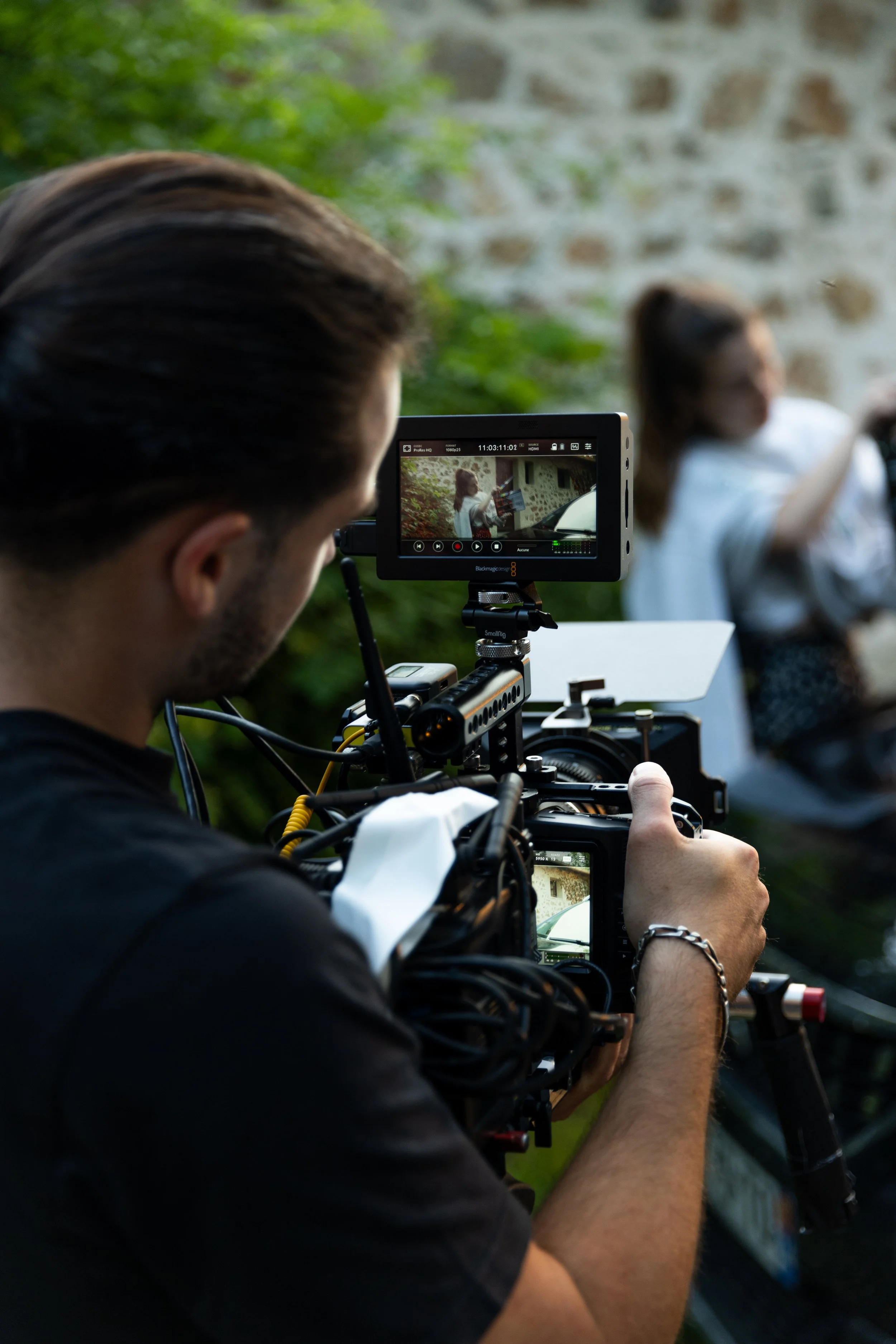 Un réalisateur filme une scène avec une caméra professionnelle, tandis qu'une femme est en arrière-plan, assise et portant des lunettes.